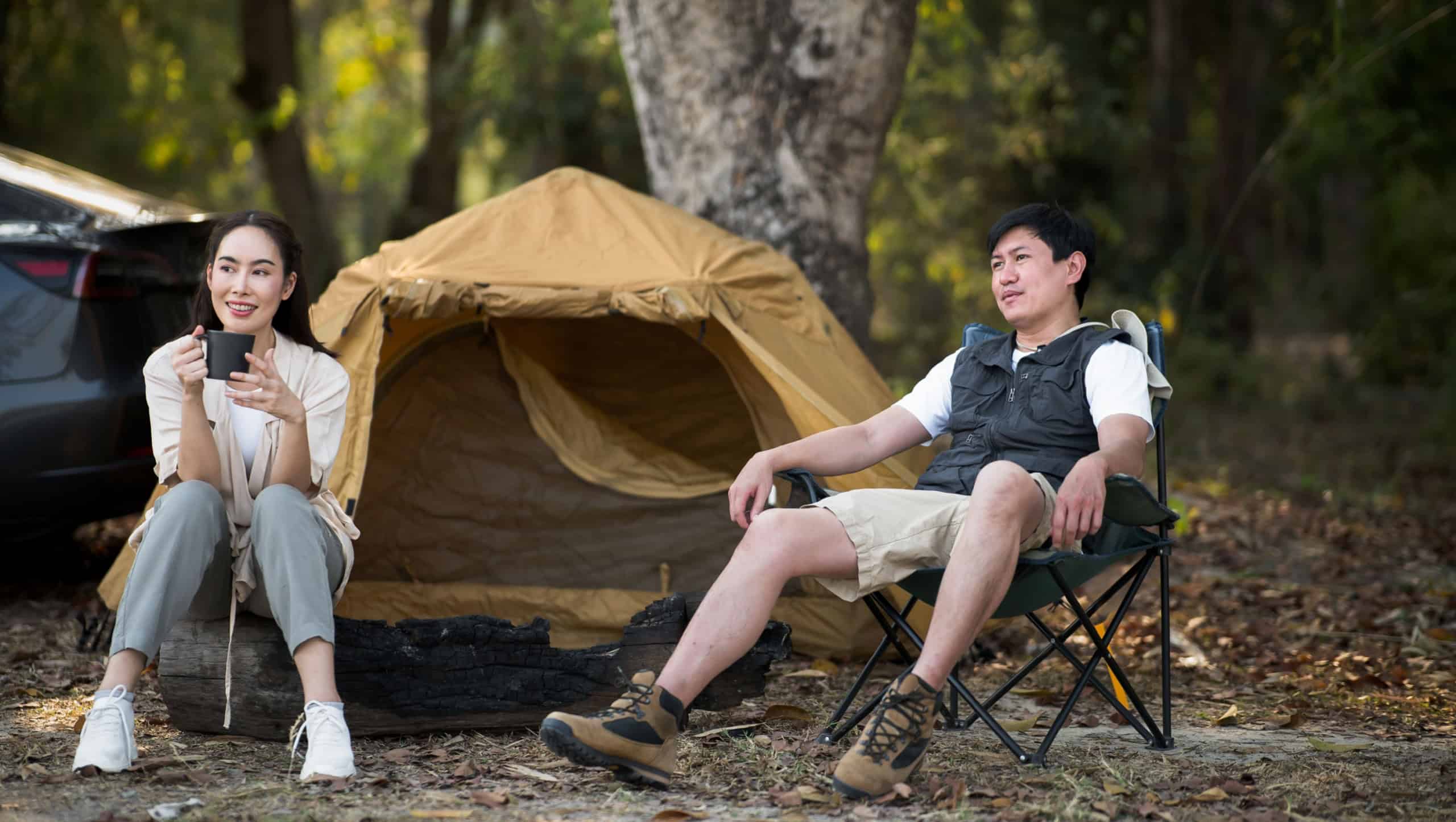A woman holding a mug and a man are sitting in front of the camping tent. Traveling together, Forest camping, Outdoor activity.