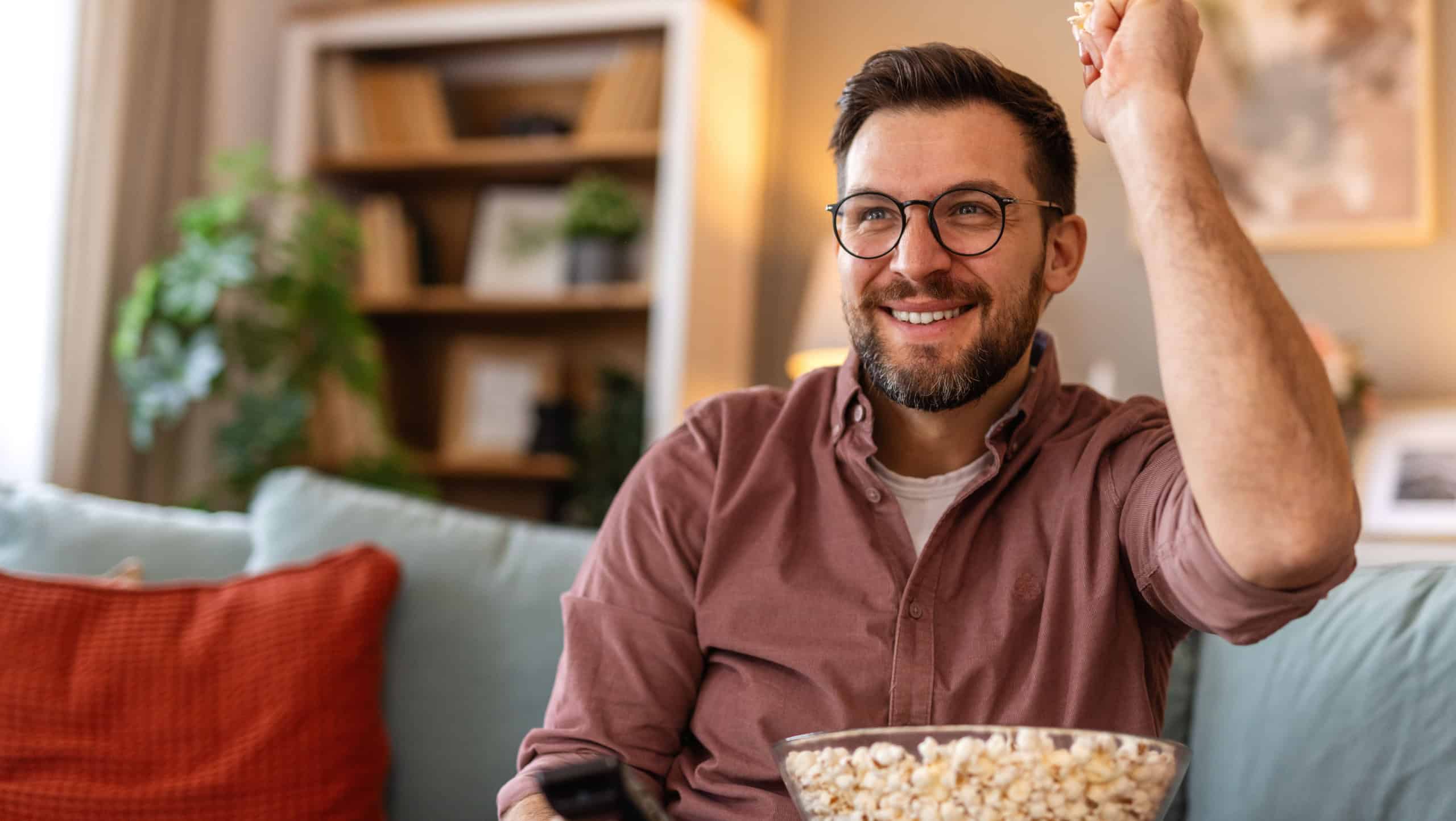A cheerful man sits on a cozy couch, holding a remote control and a big bowl of popcorn. He looks excited as he watches TV, enjoying a relaxed evening in a warmly decorated living room.