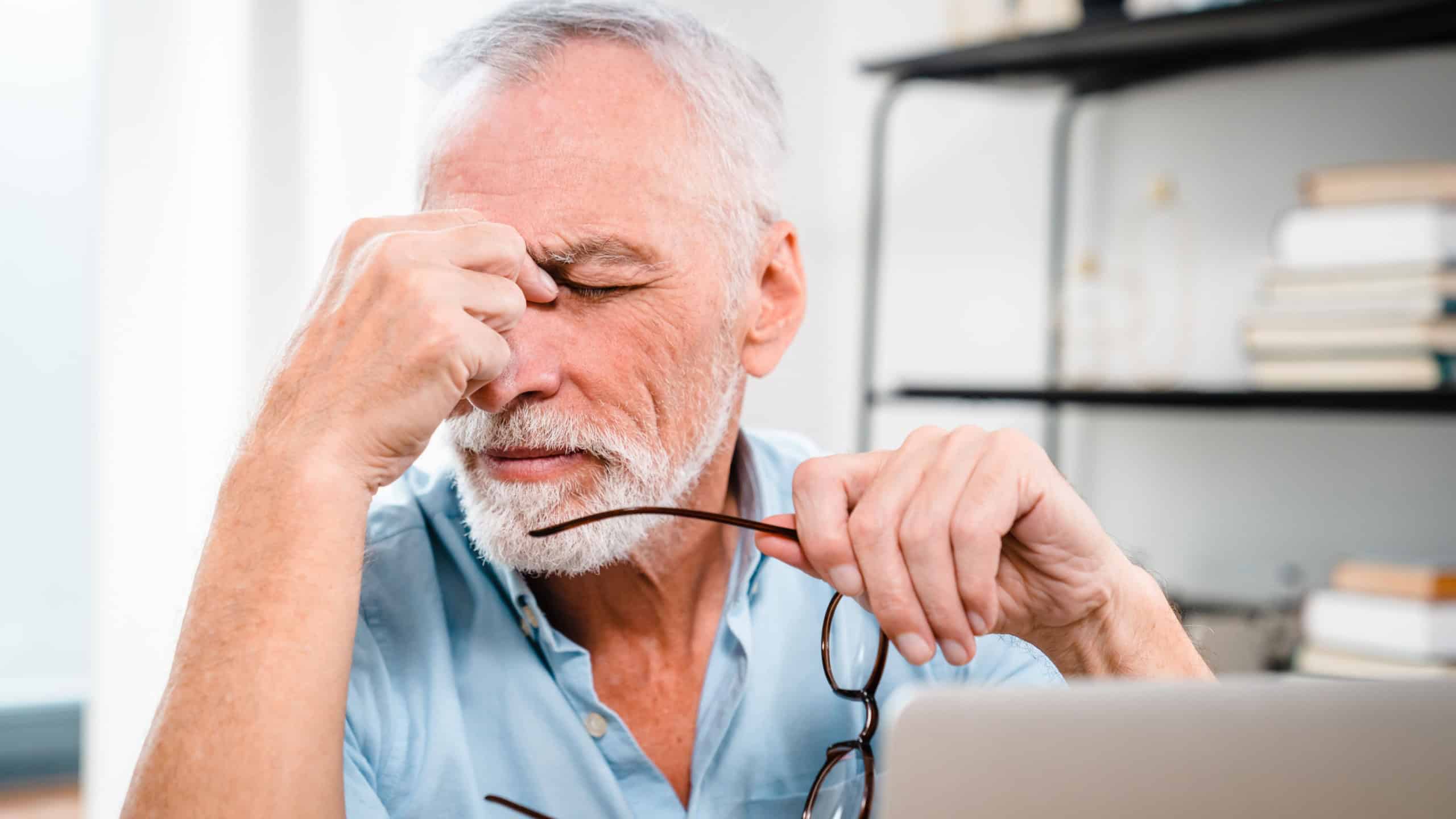 Fatigued mature old man taking off glasses suffering from tired dry irritated eyes after long computer use, senior middle aged male feels eye strain problem or blurry vision working on laptop at home
