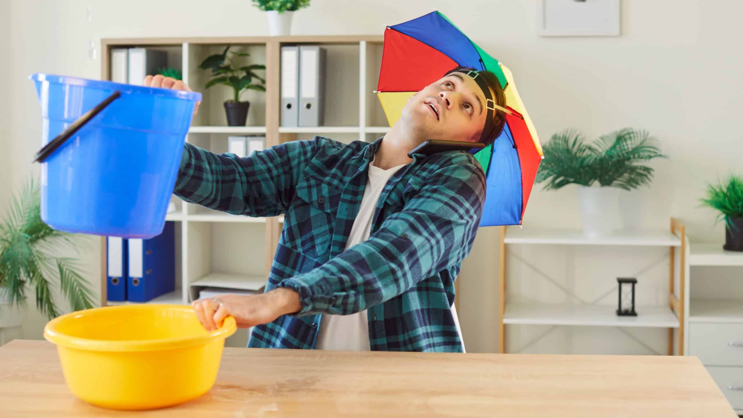 Distressed man sits at home with a bucket, calling for emergency help due to a flood, leaking roof, or broken plumbing. House damage, water problems, and urgent ceiling repair needs.