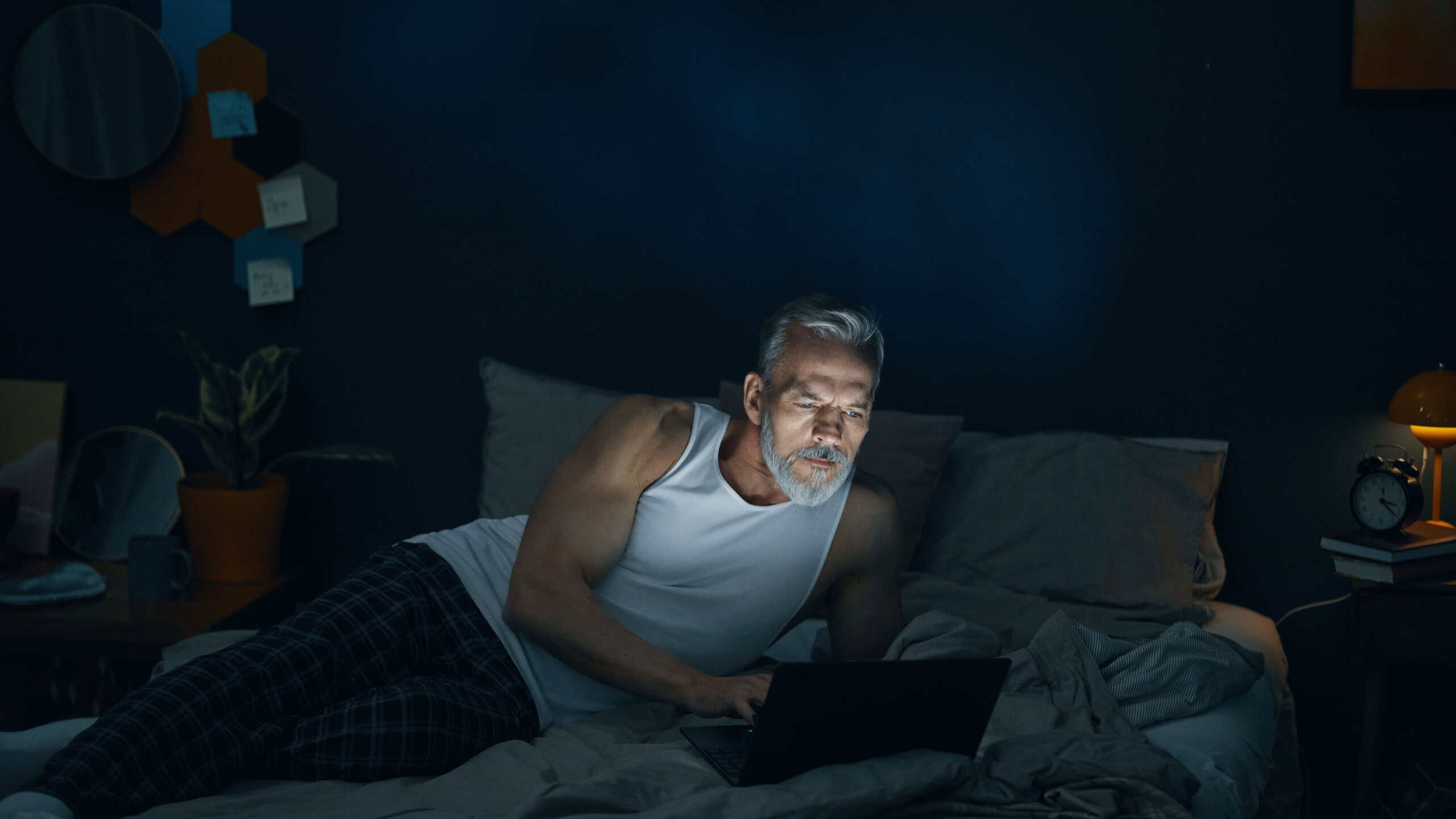 Senior man sitting in bed, illuminated by laptop screen, deeply engrossed in his work while surrounded by a dark room with minimal lighting and modern decor