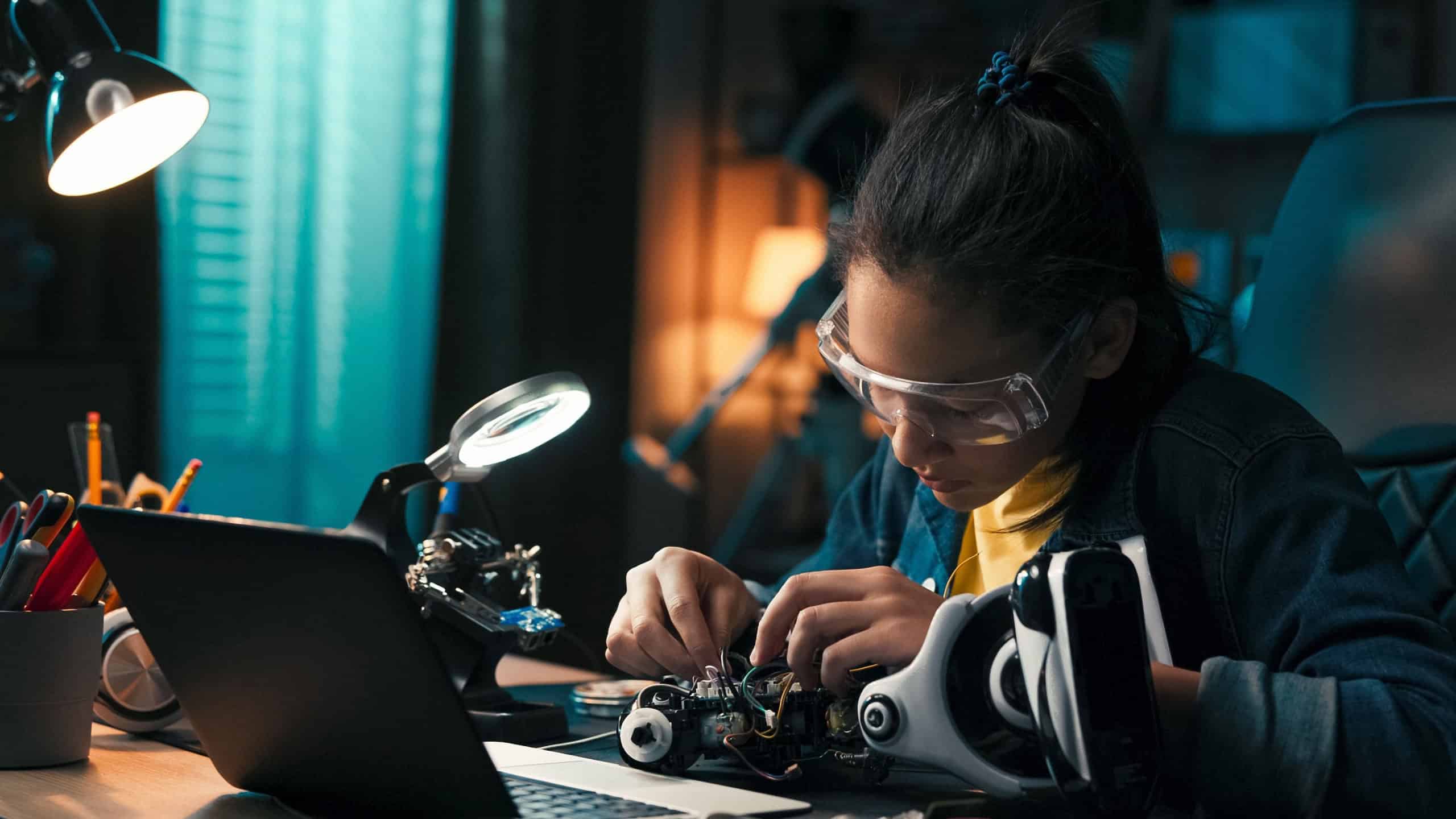 A teenage girl concentrates on robot construction at her desk in a dark room. She connects wires step by step, creating a power system for her toy using basic electronic components.
