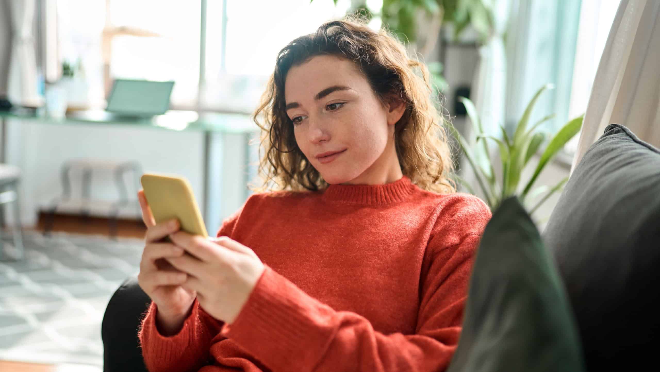 Relaxed young woman sitting on couch using cell phone technology, happy lady holding smartphone, scrolling, looking at cellphone enjoying doing online ecommerce shopping chatting in mobile ai apps.