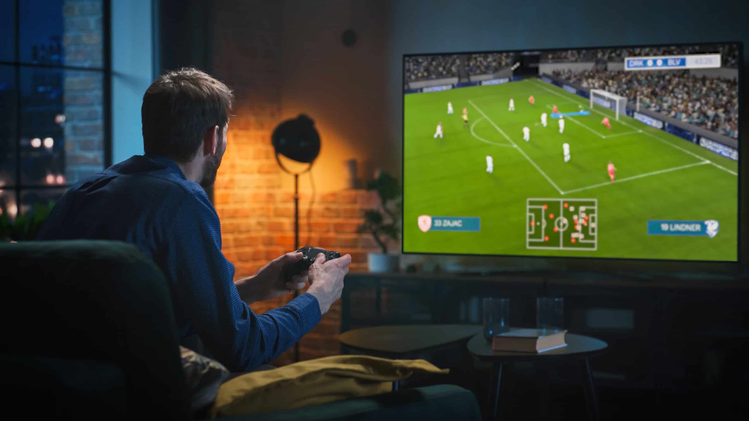 Caucasian Man Spending Time at Home in the Evening, Playing a Soccer Video Game on a Big TV Set at Home. Young Male Sitting on a Sofa, Using a Controller to Defeat a Rival Football Team Online