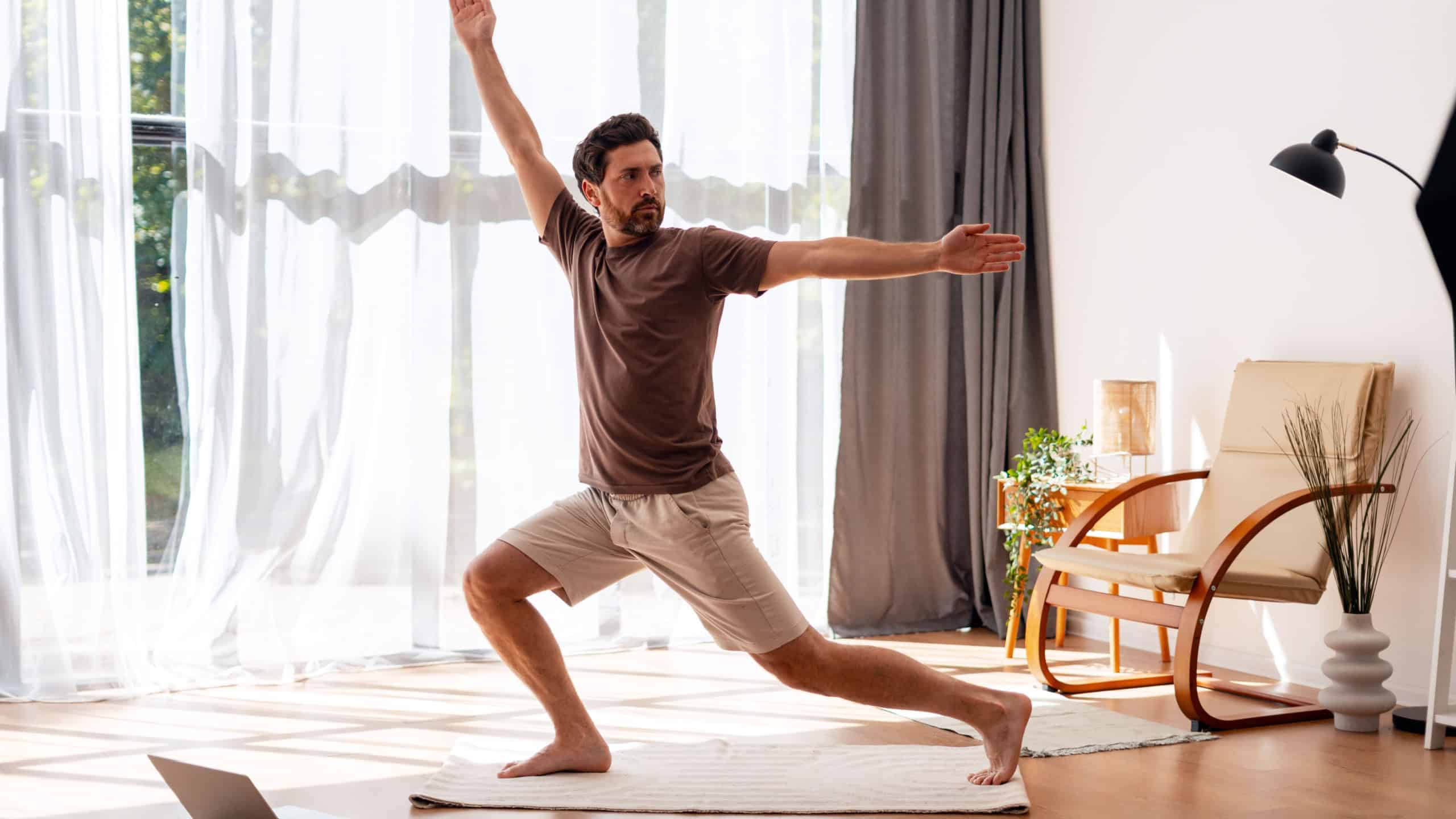 Bearded man practicing yoga warrior pose at home, using laptop for online class in his living room