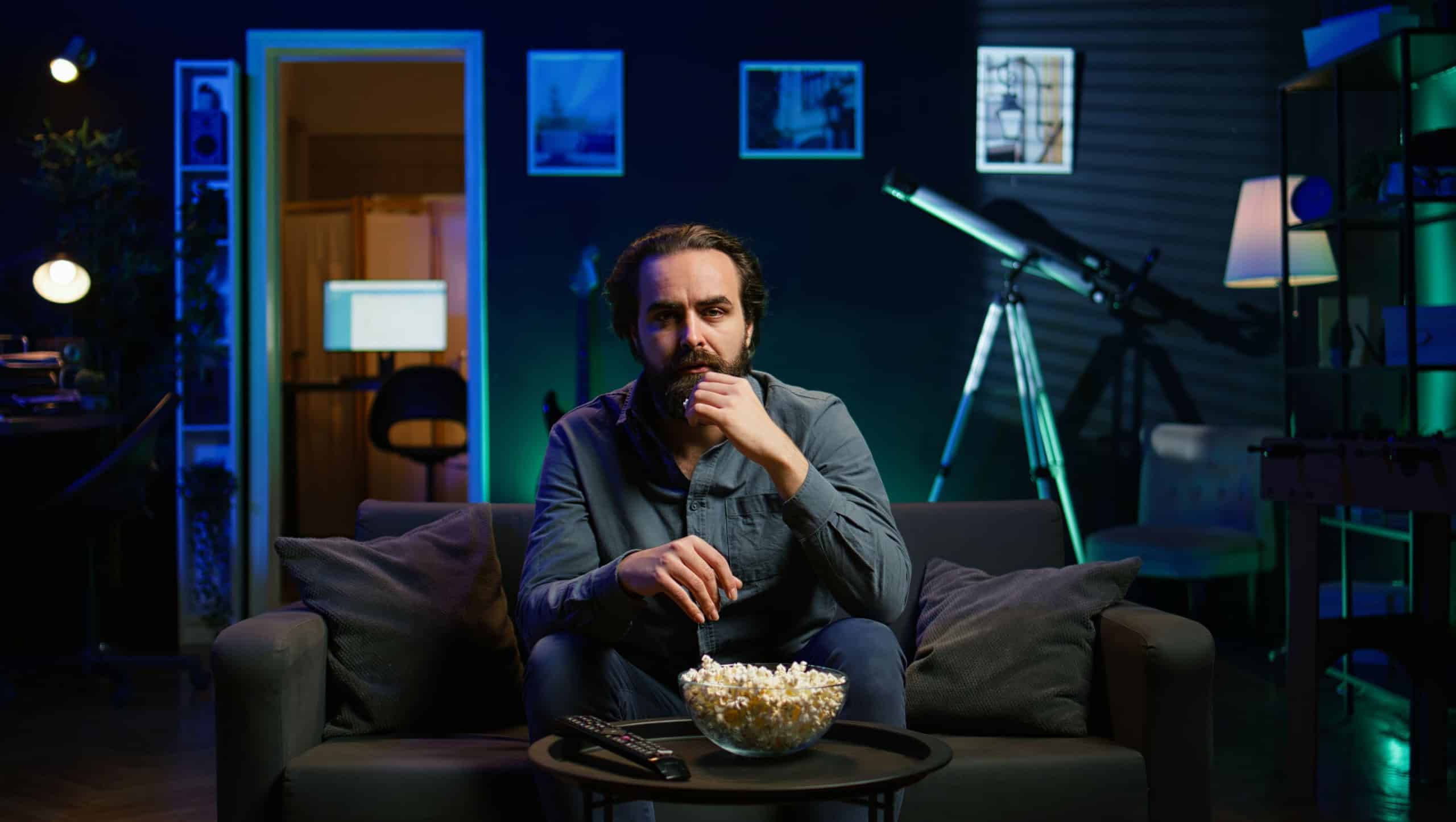 Lazy couch potato person mindlessly watching shows on TV and eating popcorn, paying close attention to screen. Sedentary man devouring snacks while consuming mindless entertainment, camera B
