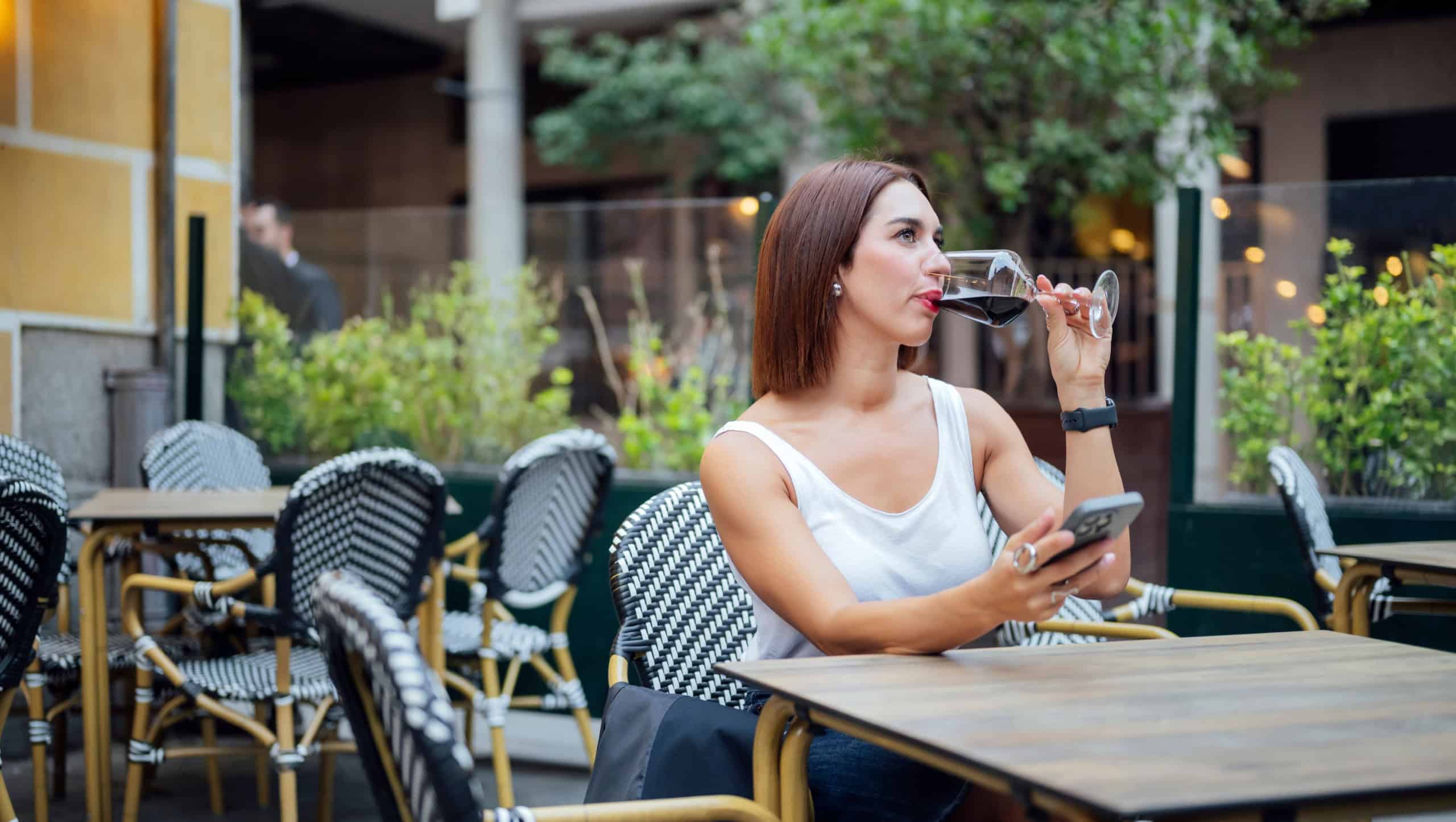 Woman relaxing in an outdoor café, holding a smartphone and enjoying a glass of red wine.