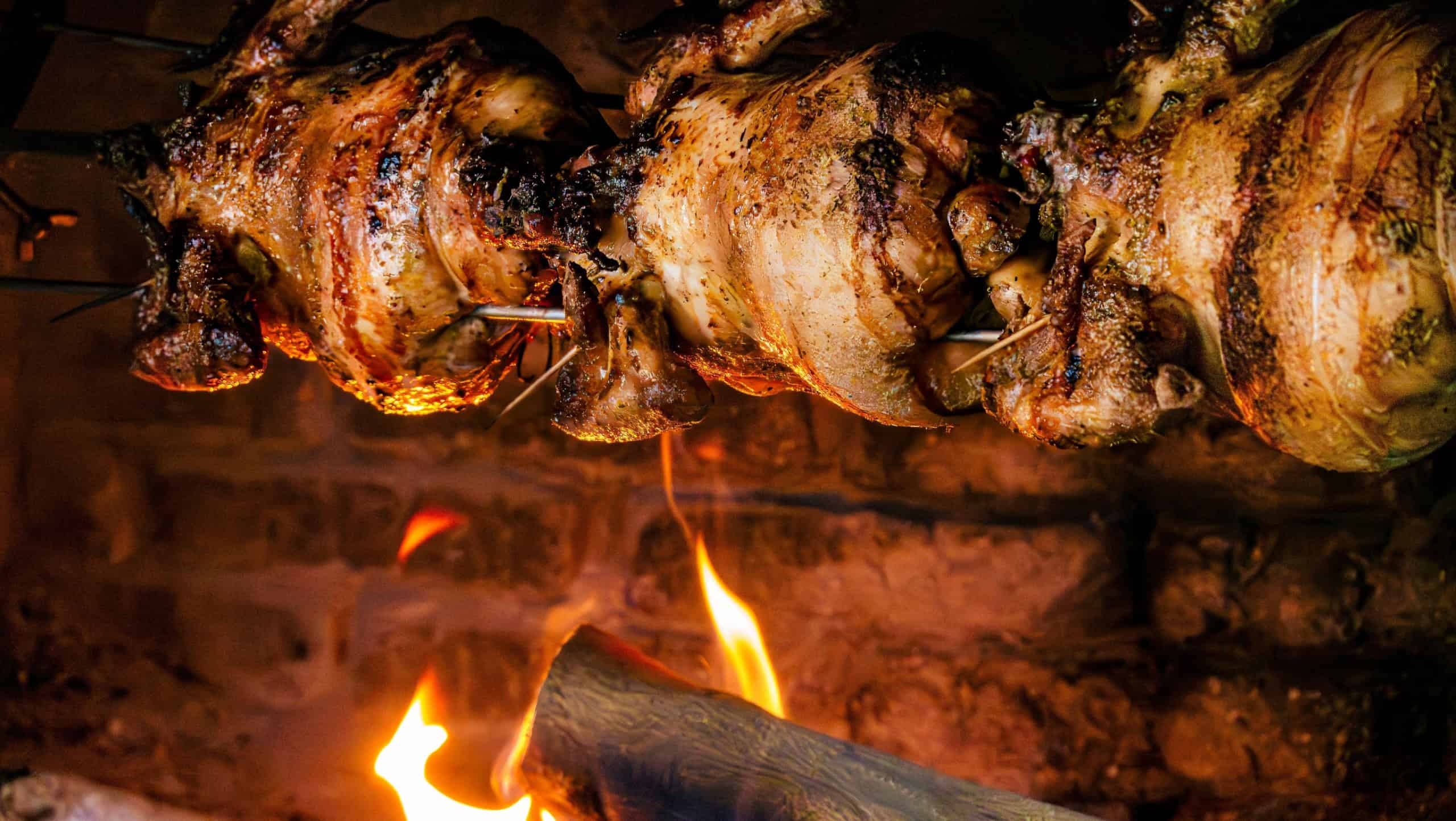 A close-up of whole chickens roasting on a rotisserie spit over an open wood fire in a traditional brick oven.