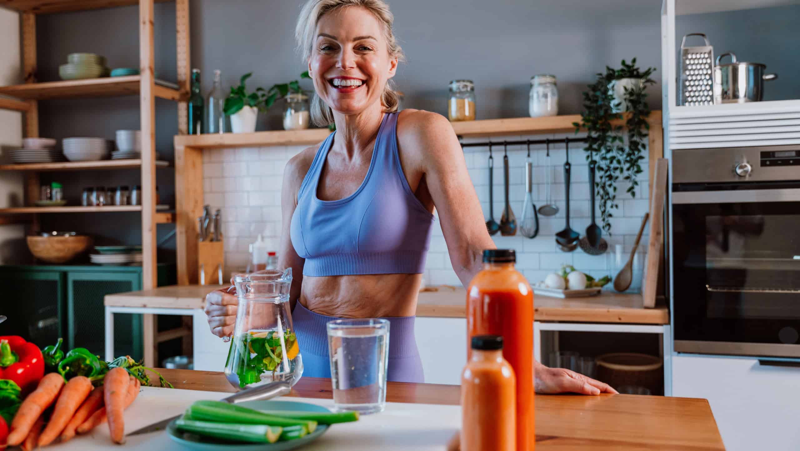 Fit mature woman wearing sportswear is pouring infused detox water into glass in kitchen at home, with fresh vegetables and healthy juices on table, promoting healthy lifestyle and wellness
