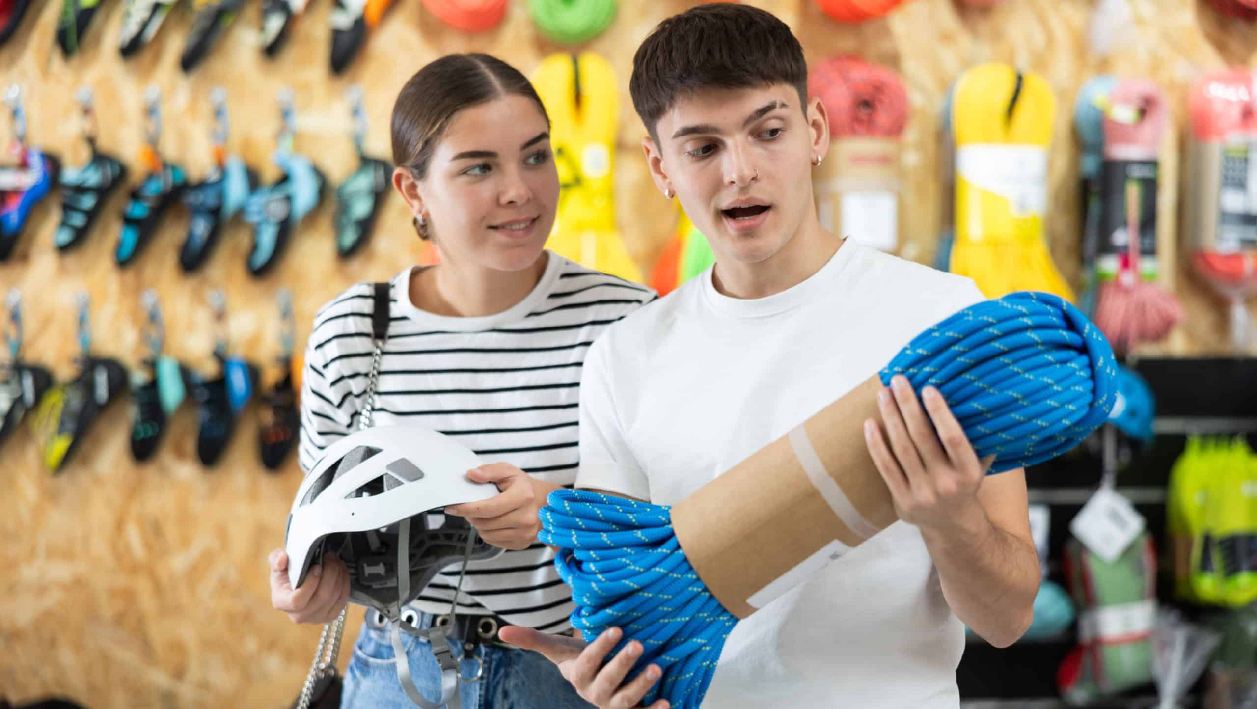 Young husband and wife choose necessary equipment for hike in tourist store. Buyers are considering rope and helmet for sports mountain climbing