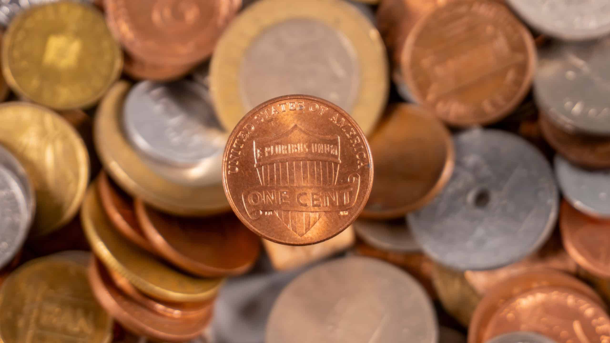 Coins of different sizes and colors are placed on a flat surface. One cent coin stands out among the scattered coins, showing various designs from different currencies.