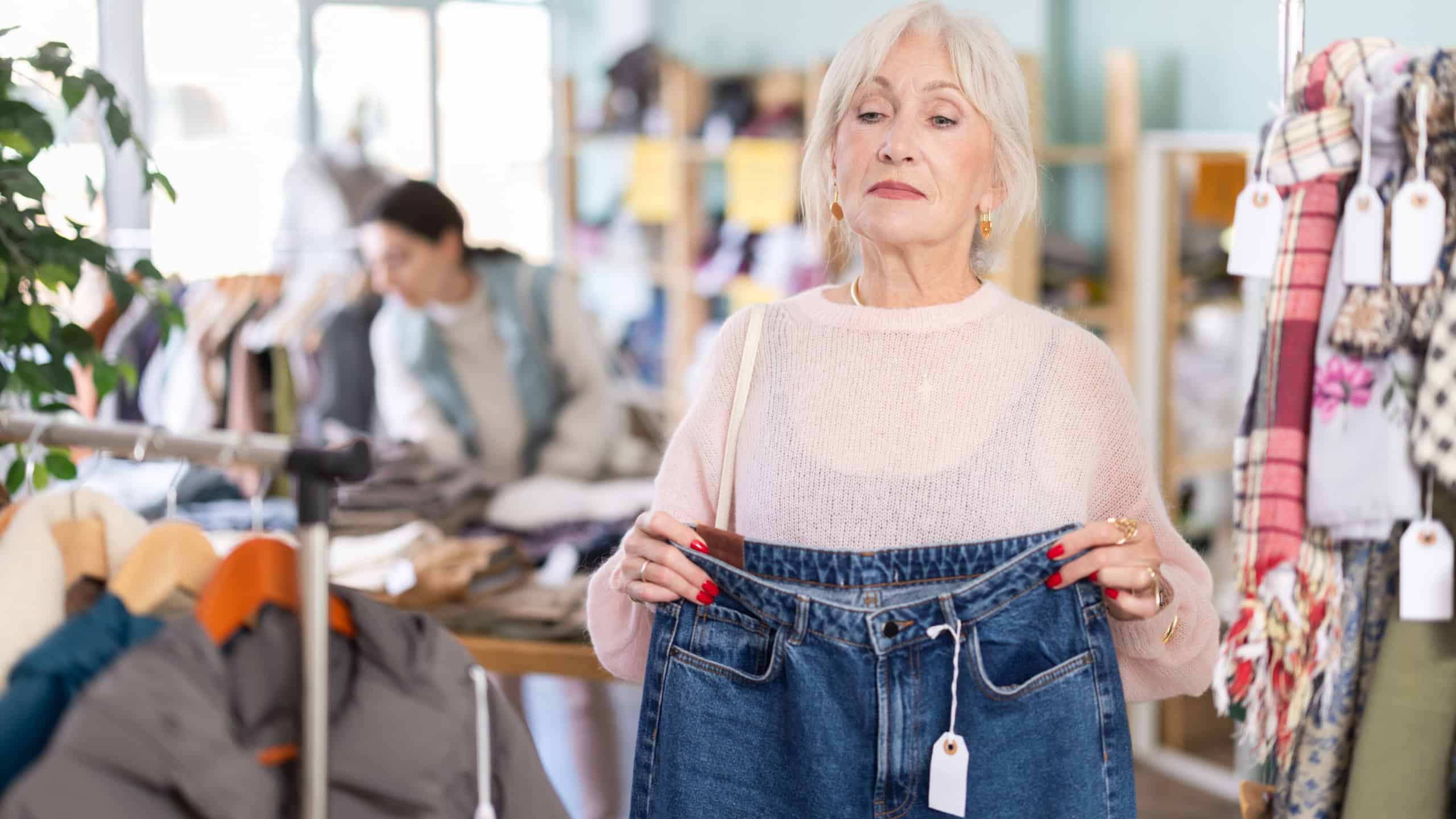 Senior woman in a store looks at stylish jeans, evaluating the size and style among the assortment of a clothing store. Elderly customer buys denim clothes