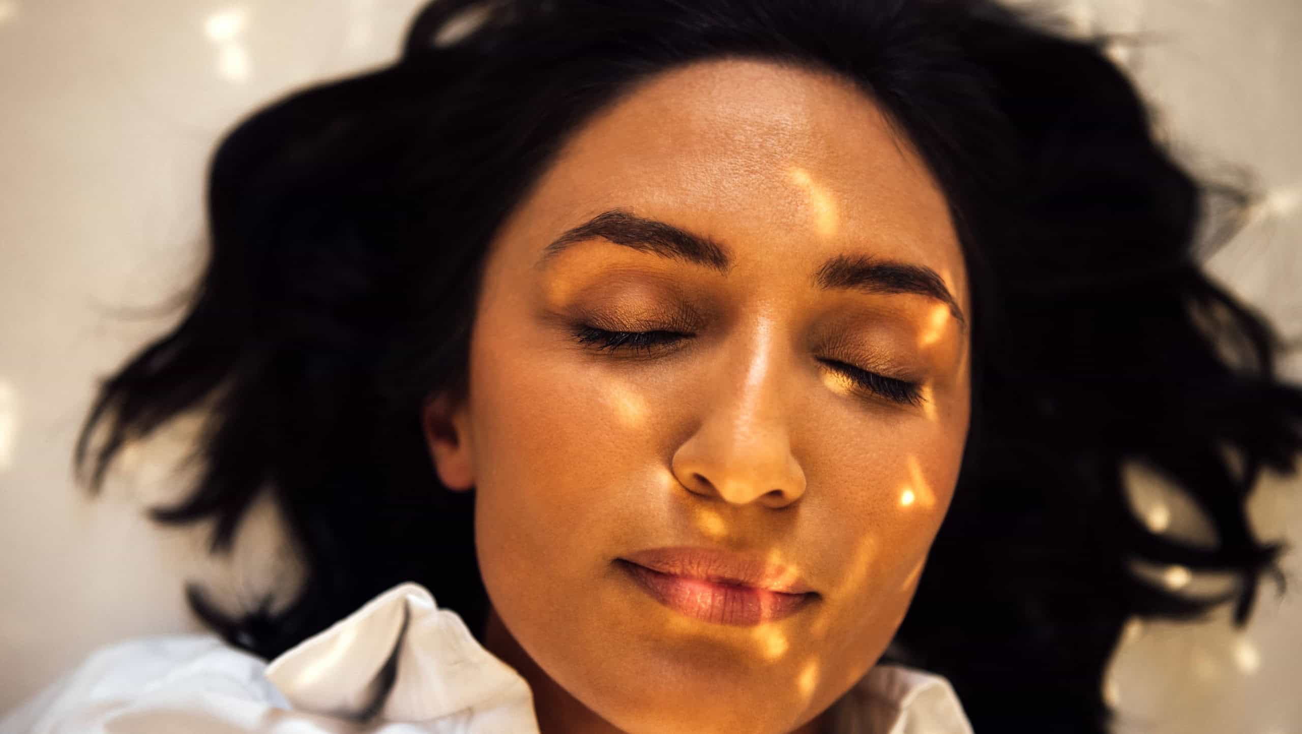 Woman with closed eyes rests on soft surface, bathed in warm sunlight, creating a tranquil ambiance, emphasizing relaxation and inner peace in a serene setting