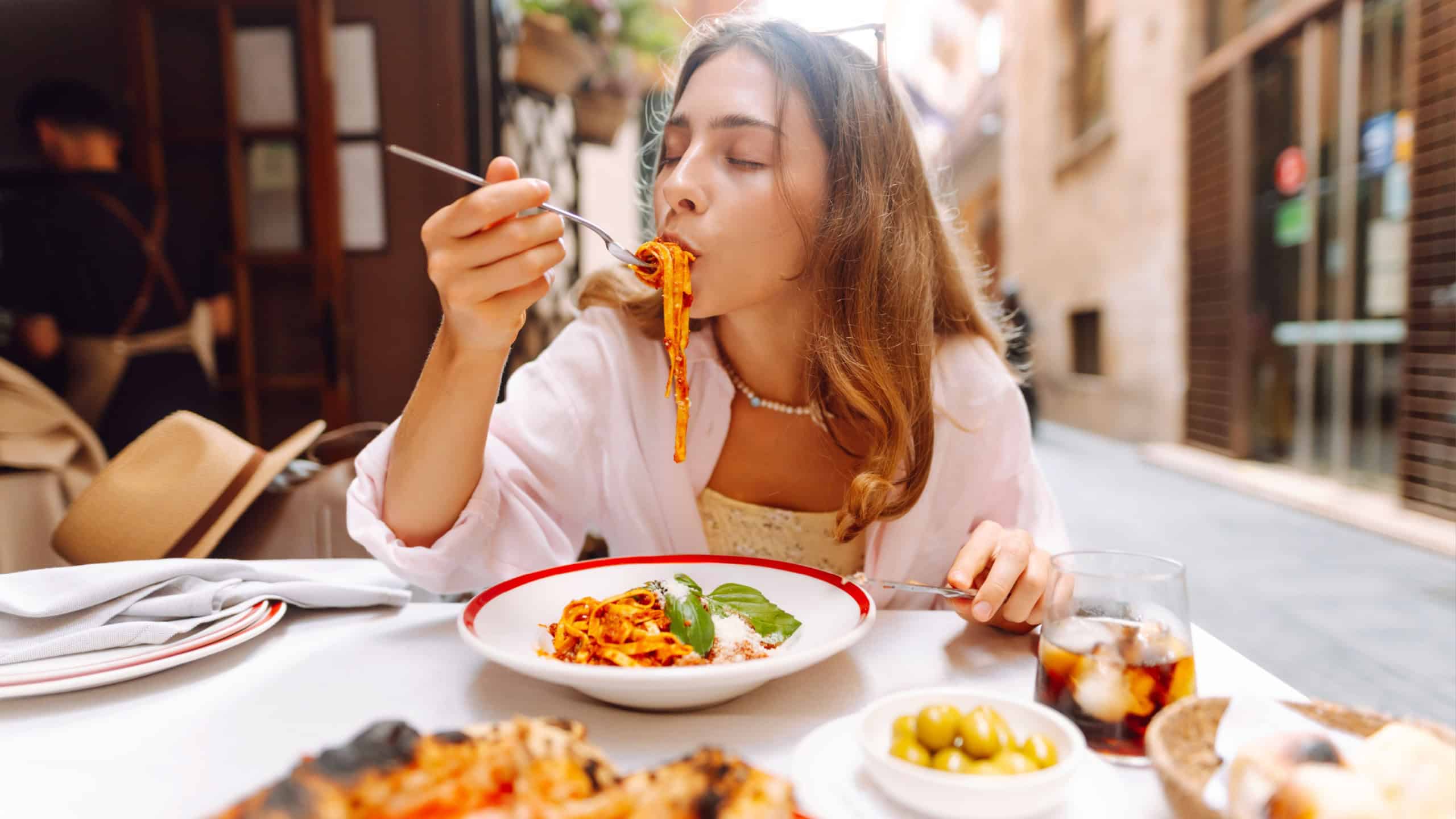 A happy woman eats fresh Italian pasta at a cafe table. A young tourist enjoys spaghetti on a sunny day on the terrace. Concept of food, enjoyment.