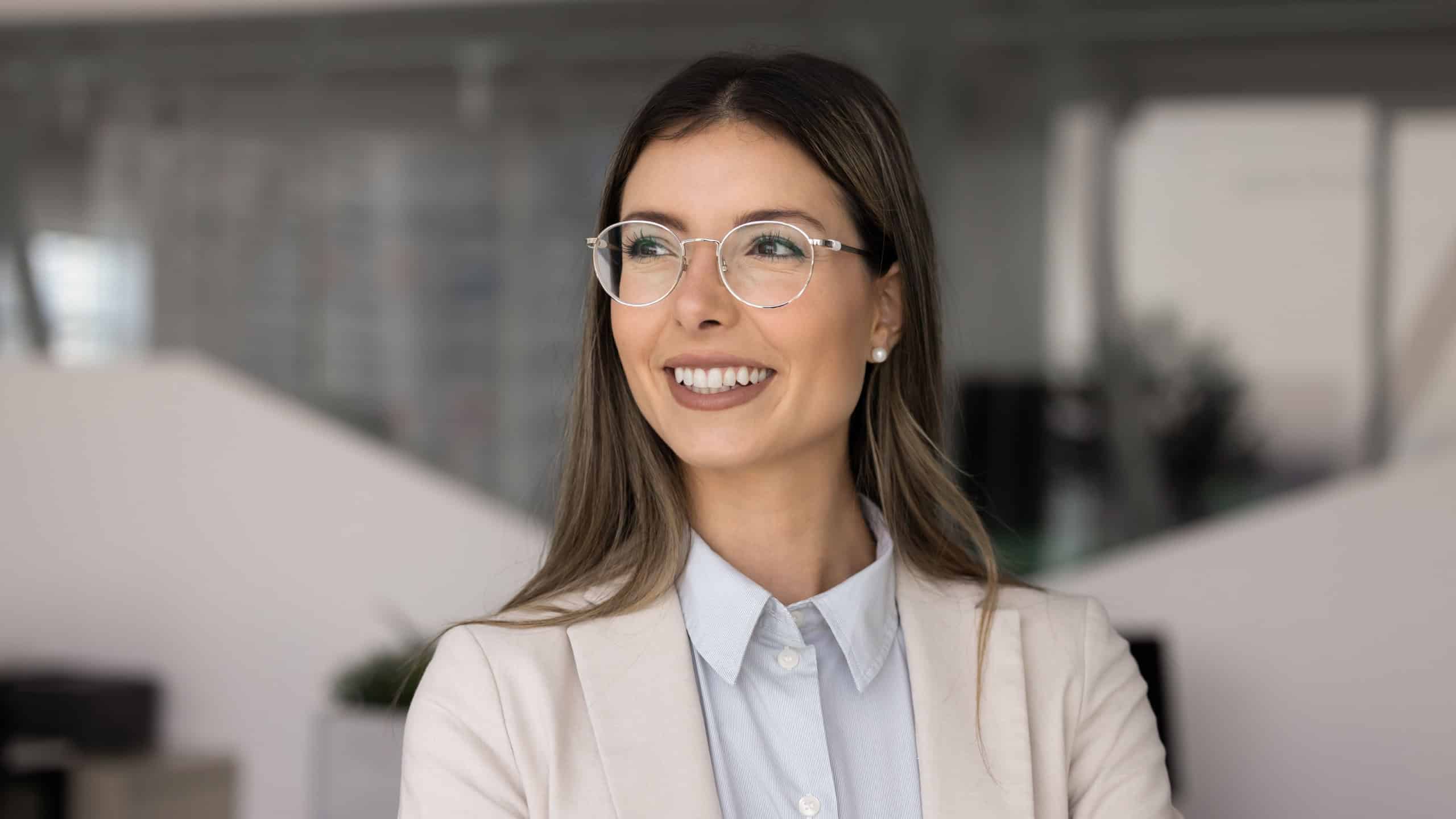 Head shot portrait of confident young Hispanic businesswoman standing alone in modern office, looks aside with contented smile, daydreaming, planning. Business vision, opportunities and career growth