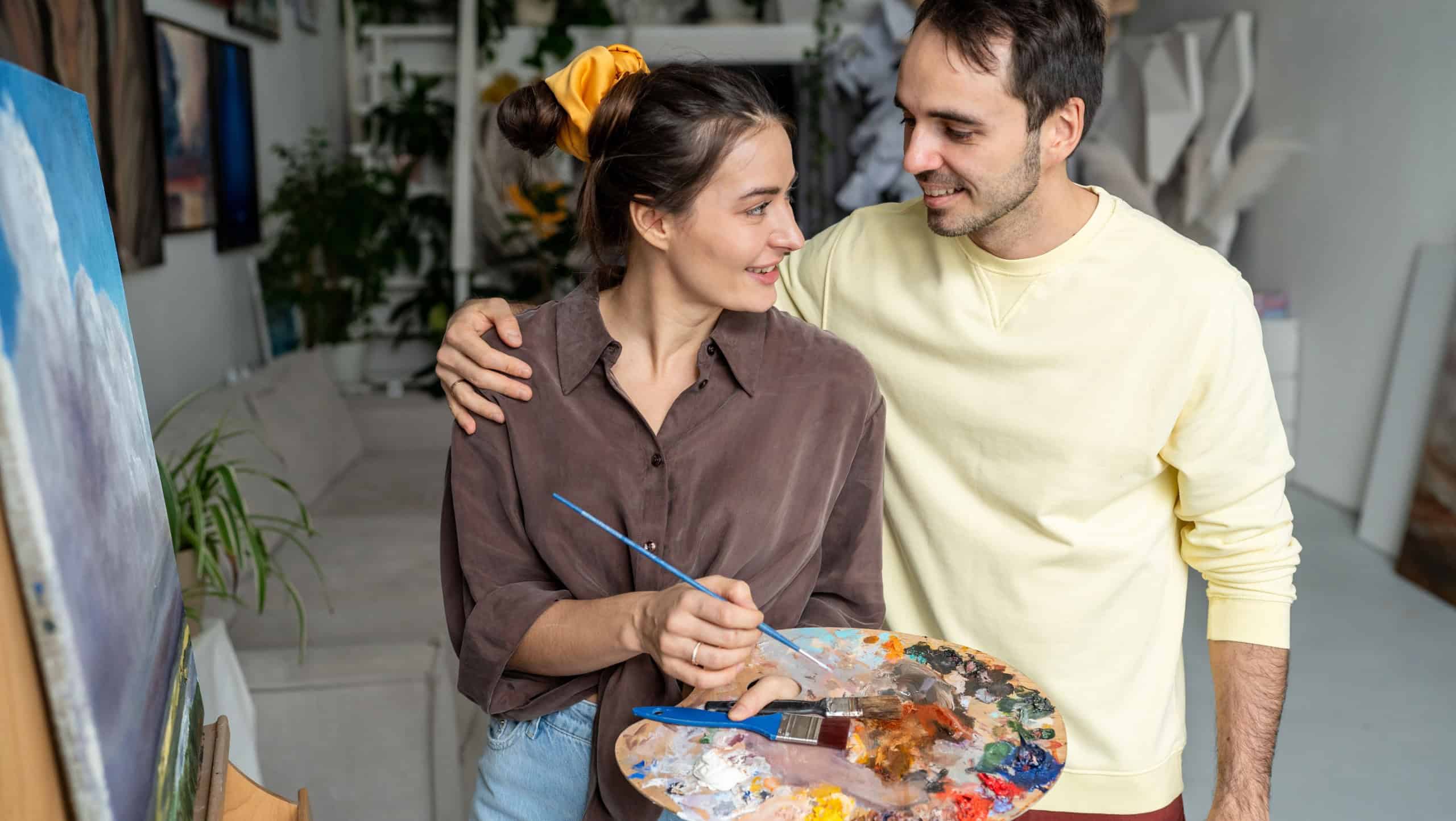 Loving couple of artists working on painting. Woman creating oil painting, curious man watches with interest leans on shoulder of beloved wife. Happy satisfied painters spouses at home studio workshop