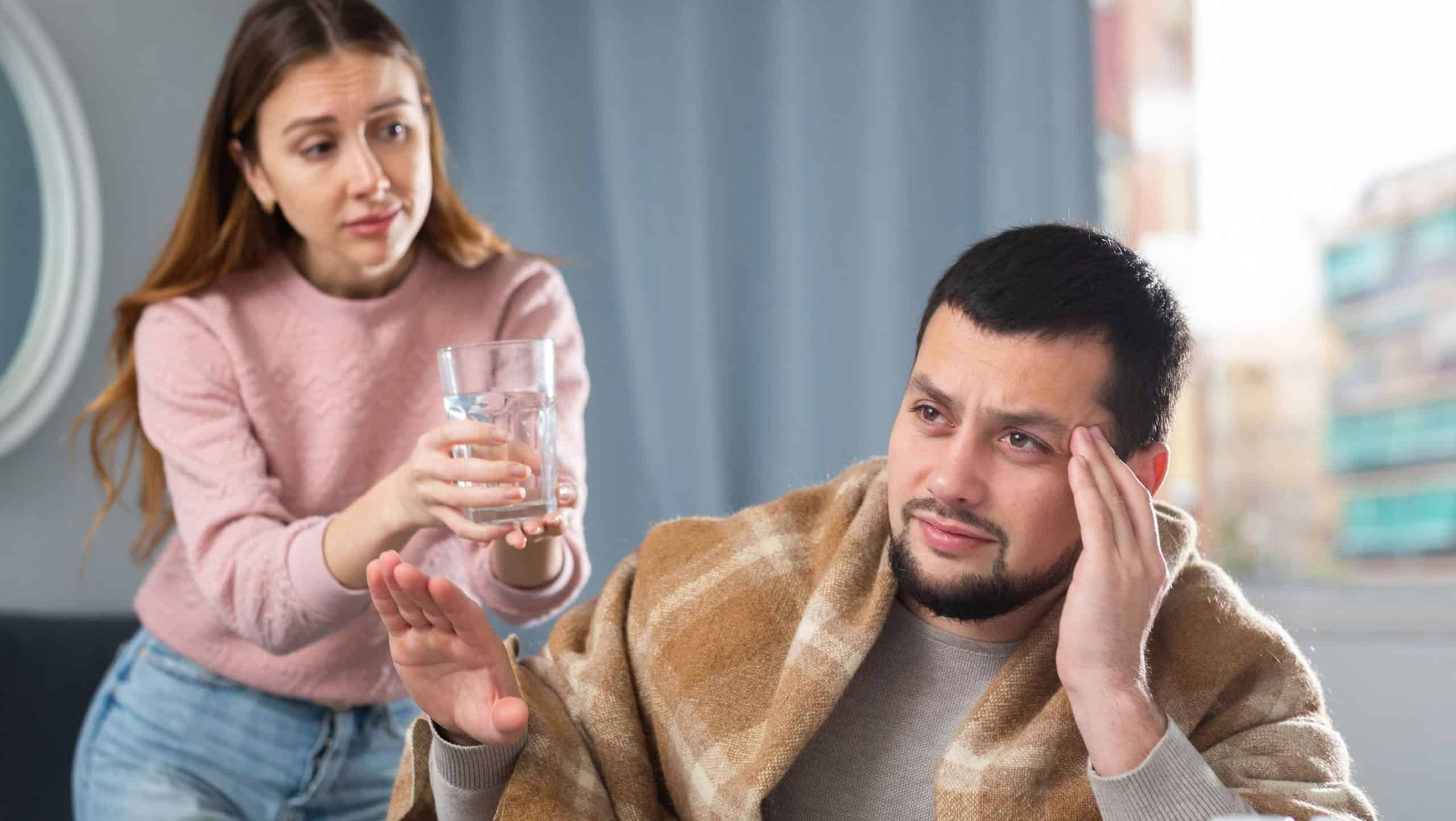Young man wrapped in blanket sitting at table at home with cold symptoms suffering from headache, refusing with gesture of water served by worried caring wife