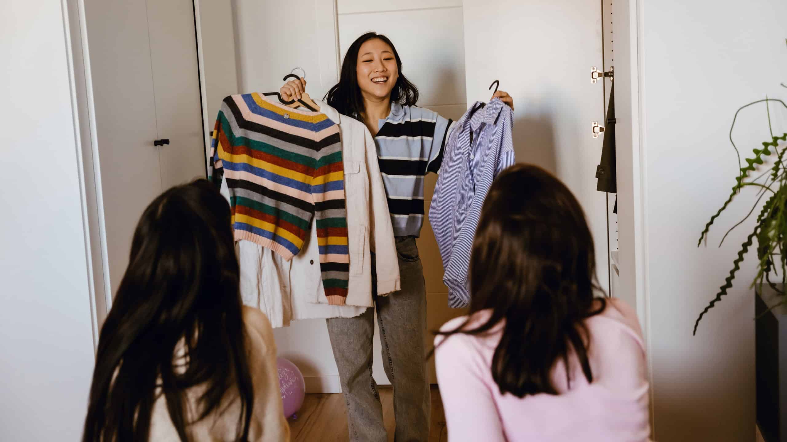 A woman stands and smiles while showing off her clothes to two female friends sitting across from her