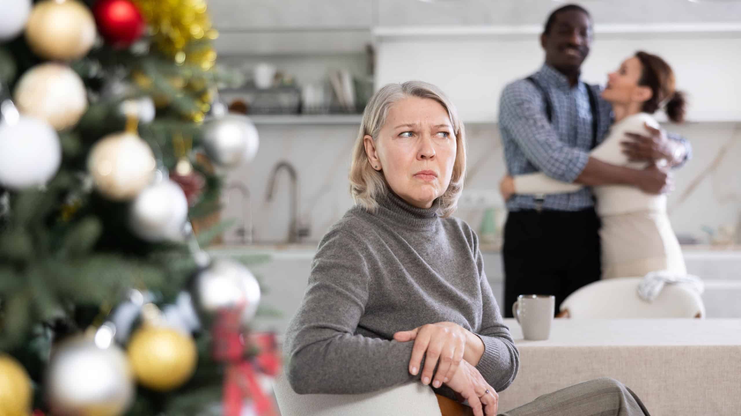Elderly woman sad while sitting at table in kitchen with happy couple of man and woman