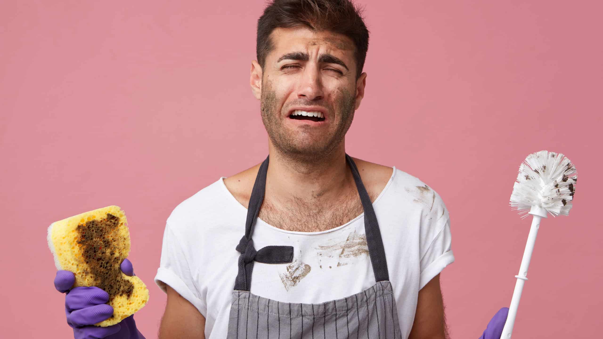 Portrait of young male doing chores having sorrorful expression not wanting to clean everything. Male wearing apron holding sponge with brush having tired and frustrated look after tidying up