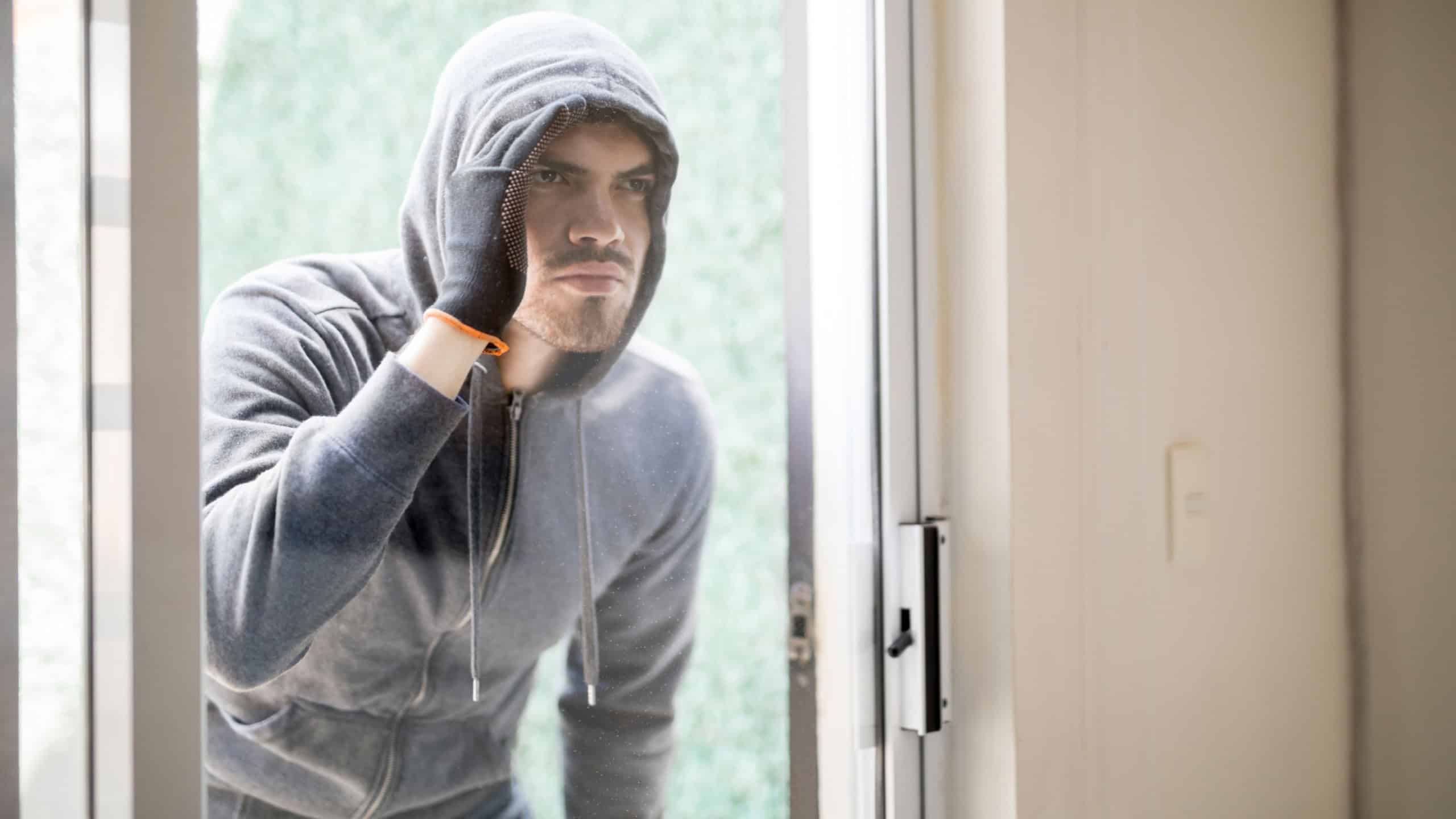 Portrait of a male robber wearing a hoodie and gloves looking inside a house through a glass door