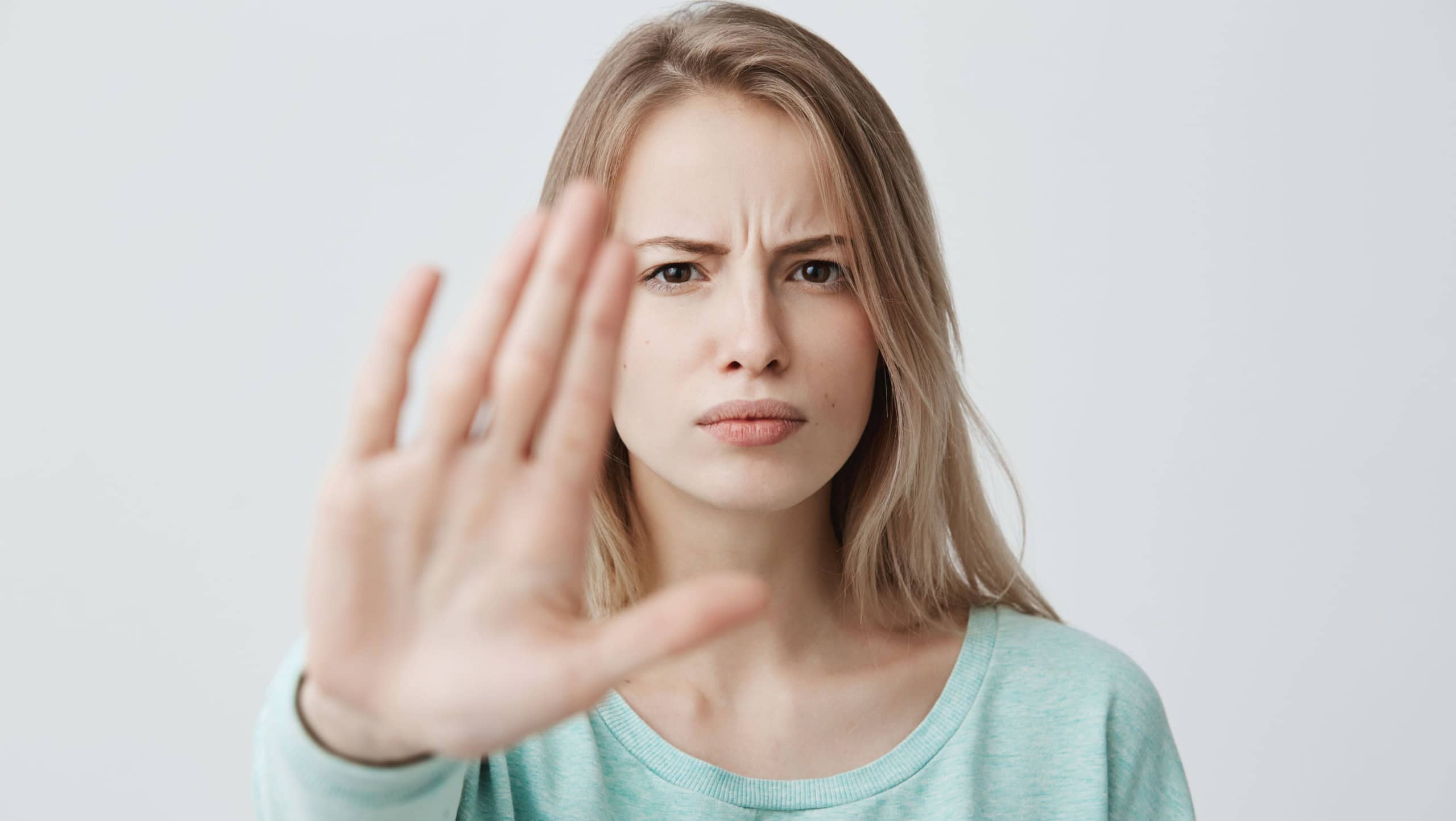 Body language. Disgusted stressed out angry blonde young female with straight fair hair posing against studio wall, keeping hand in stop gesture, trying to defend herself as if saying: Stay away from