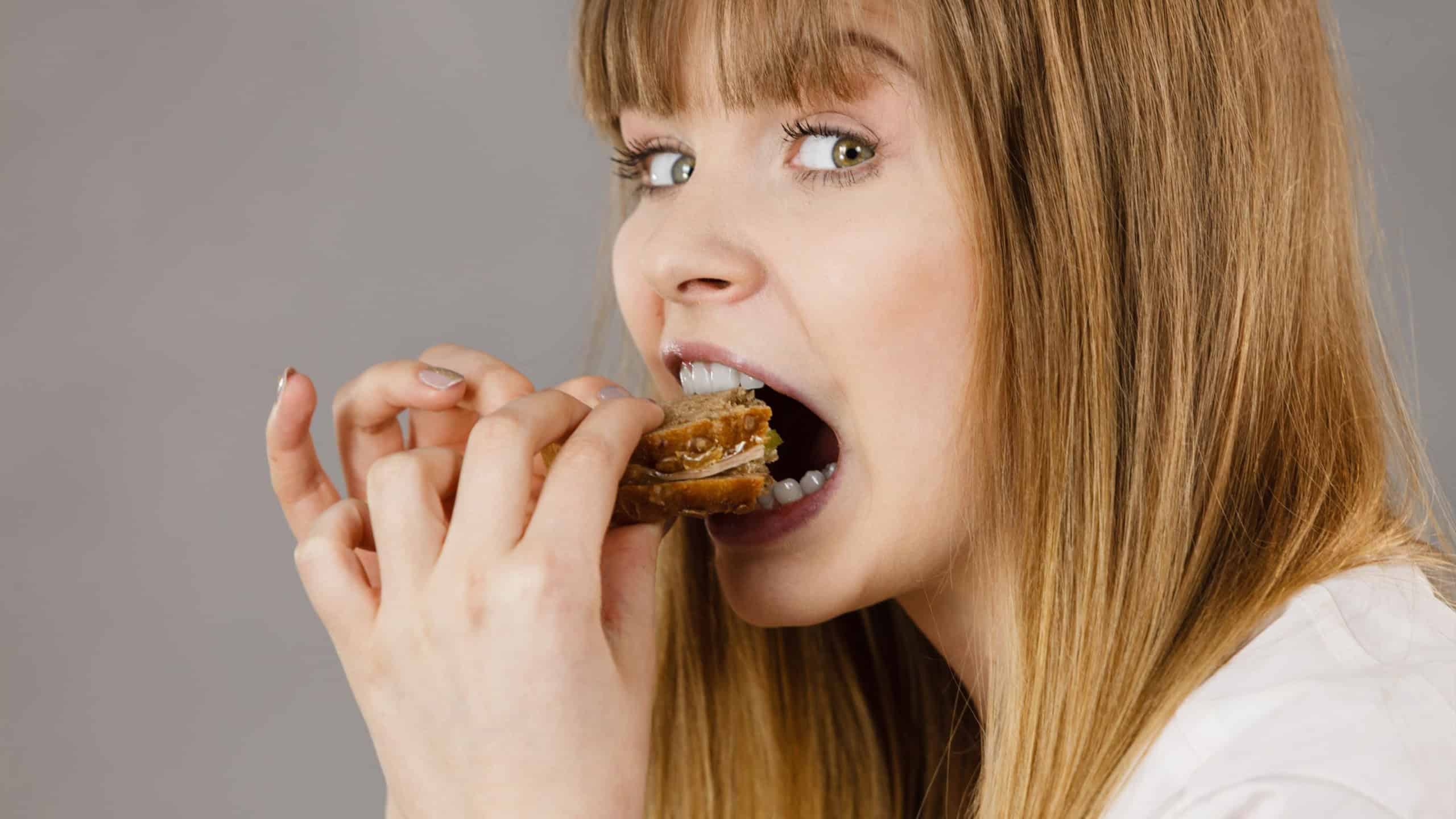 Young woman eating sandwich, taking bite with wide open mouth. Food, calories, dieting concept. Studio shot on grey background, profile view.