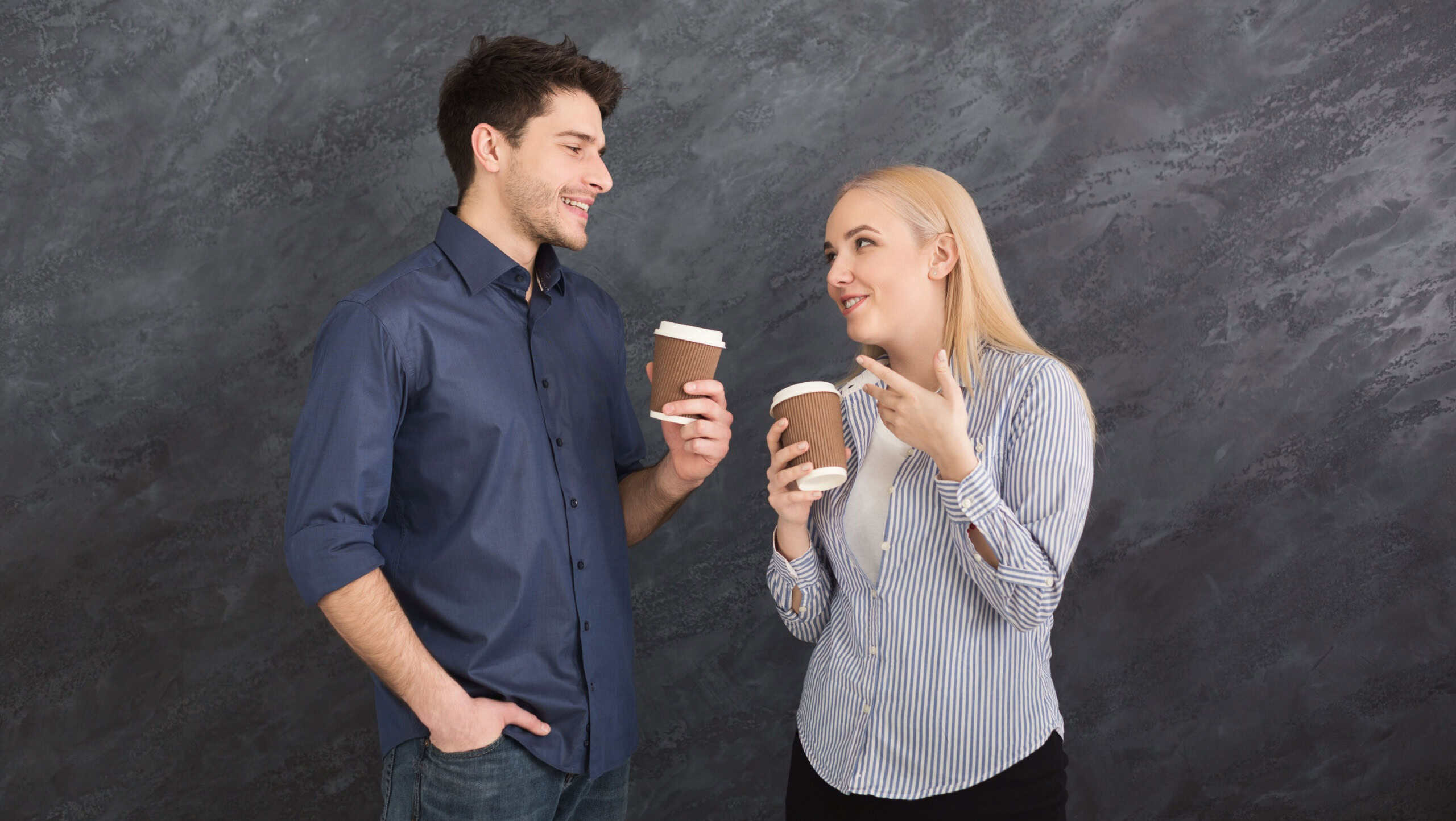 Coffee break. Couple of young colleagues drinking coffee, having nice talk, gray studio background, copy space
