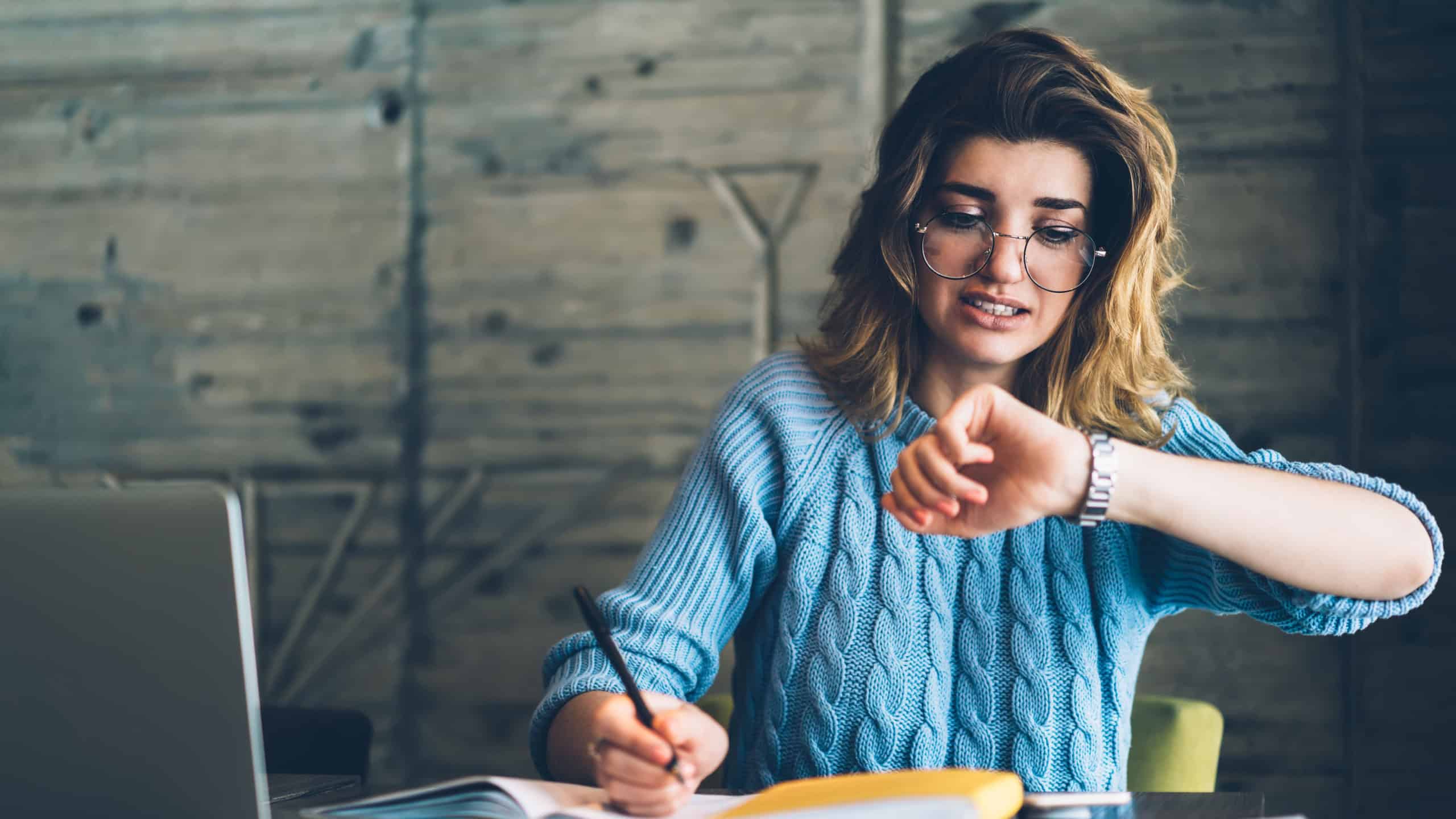 Caucasian hipster girl checking time on wristwatch feeling unhappy and hurrying up on deadline learning and doing homework using literature and laptop at wifi zone, stressed student preparing for exam