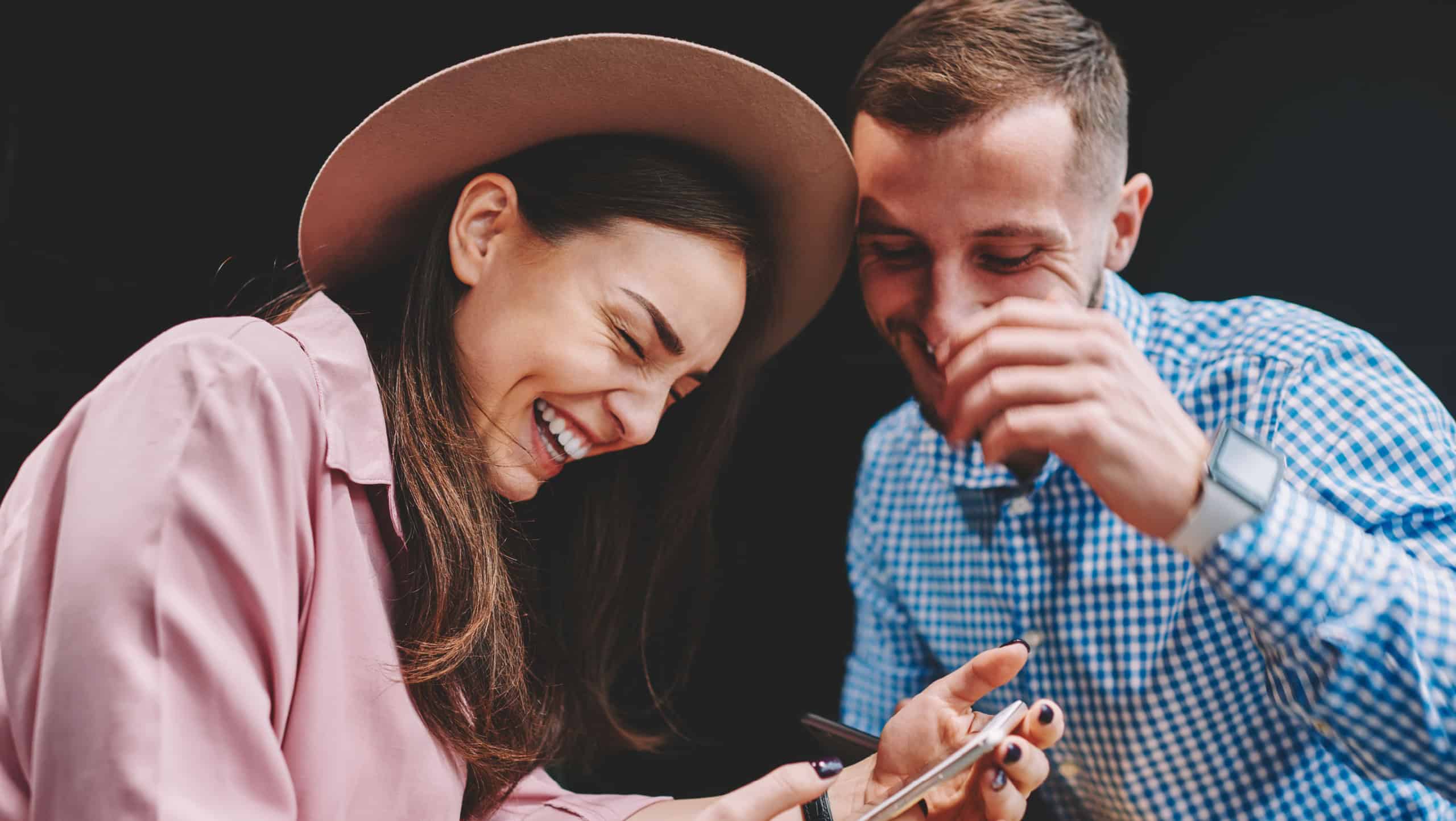 Happy couple in love laughing while watching funny video on smartphone device using free 4G internet connection.Positive girlfriend and boyfriend having fun during messaging with friends on cellular