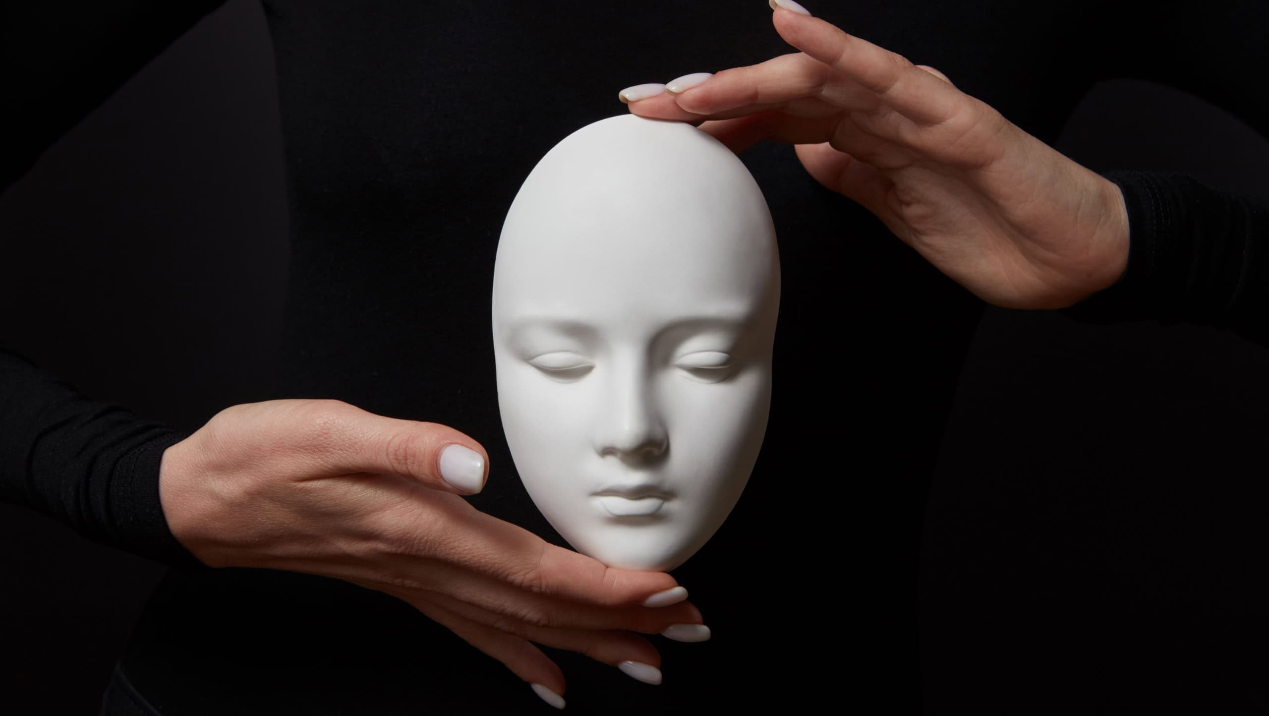 White plaster mask face is holding woman's fingers on a black background, copy space. Concept social psychological masks