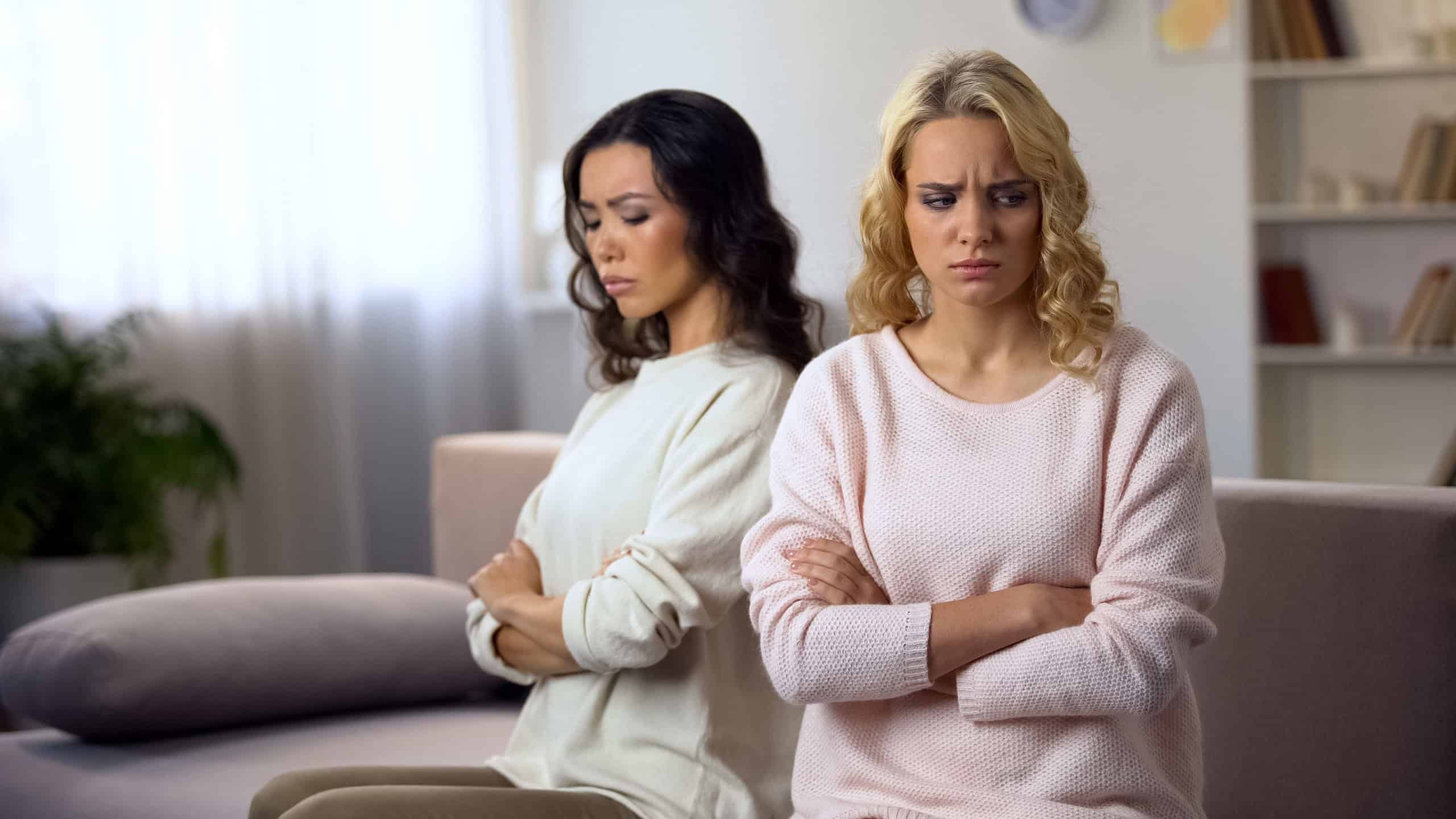 Two female friends sitting separately on sofa at home, offended after quarrel