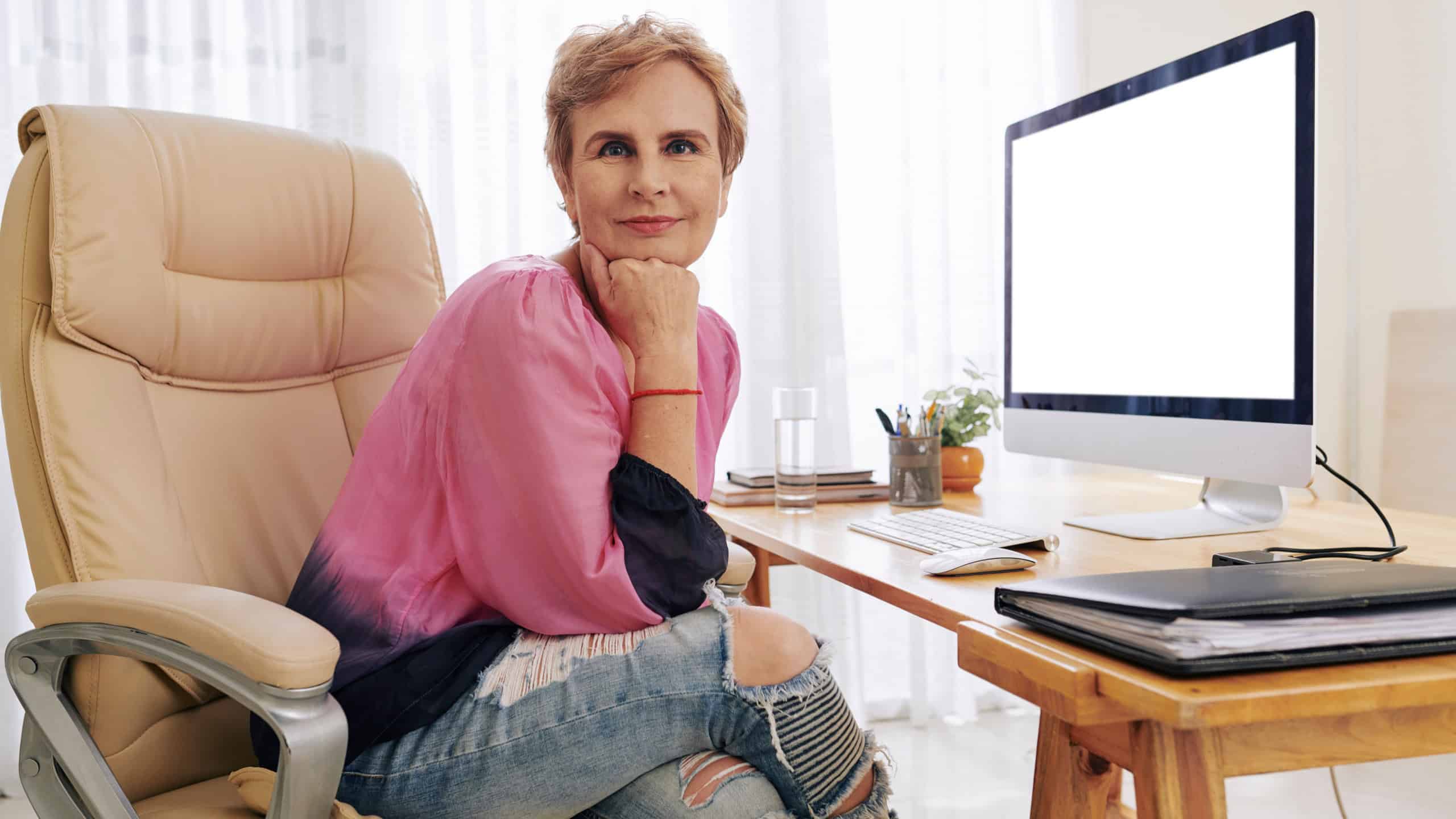 Positive successful business lady sitting in office chair in gradient blouse and ripped jeans