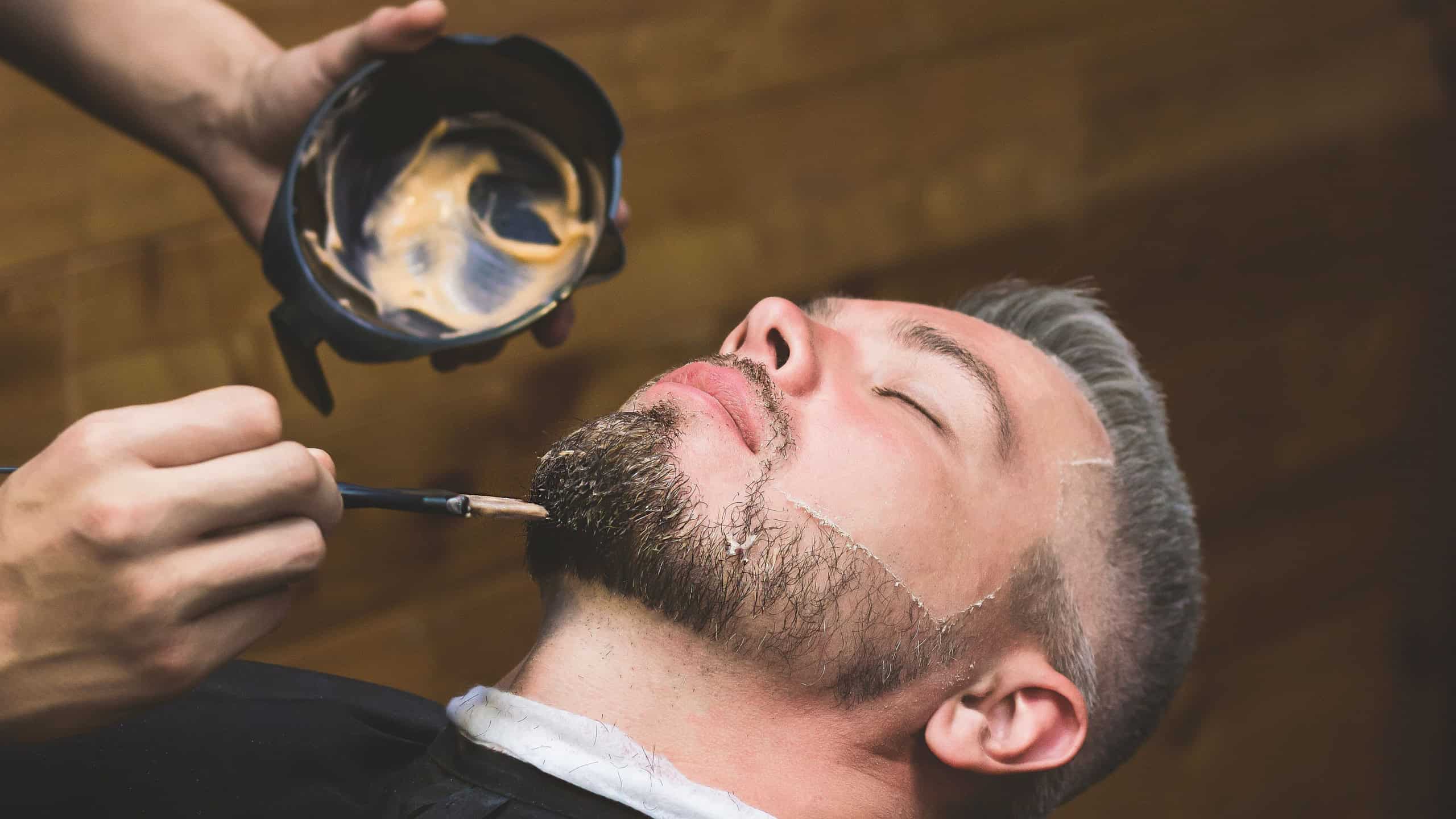 Hairdresser paints a beard to a man, male in a beauty studio. Close-up face and person, beard and hair in a barbershop. Handsome boy hipster caring for his looks.