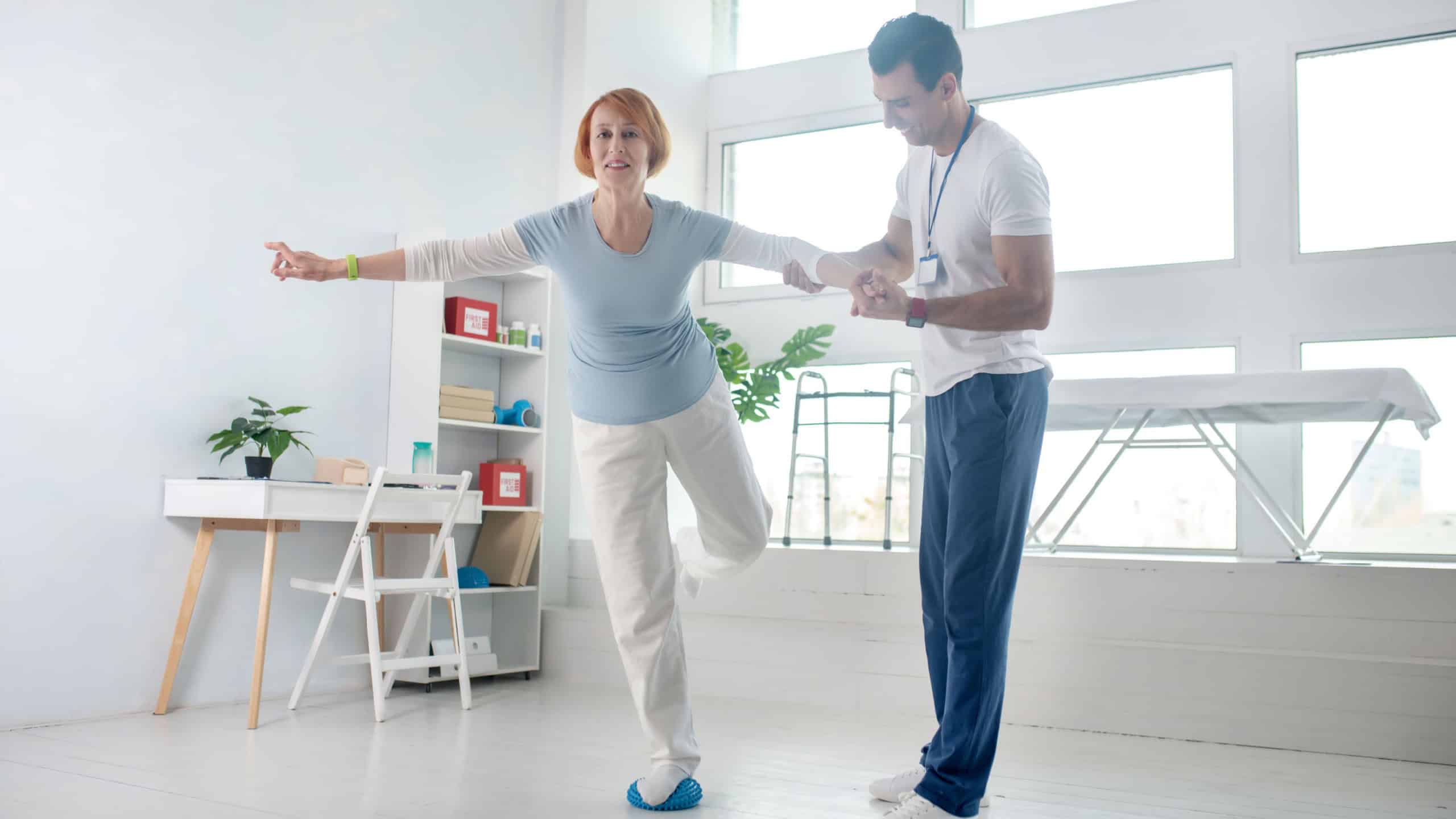 Balance exercise. Positive aged woman standing on one leg while holding the hand of her instructor