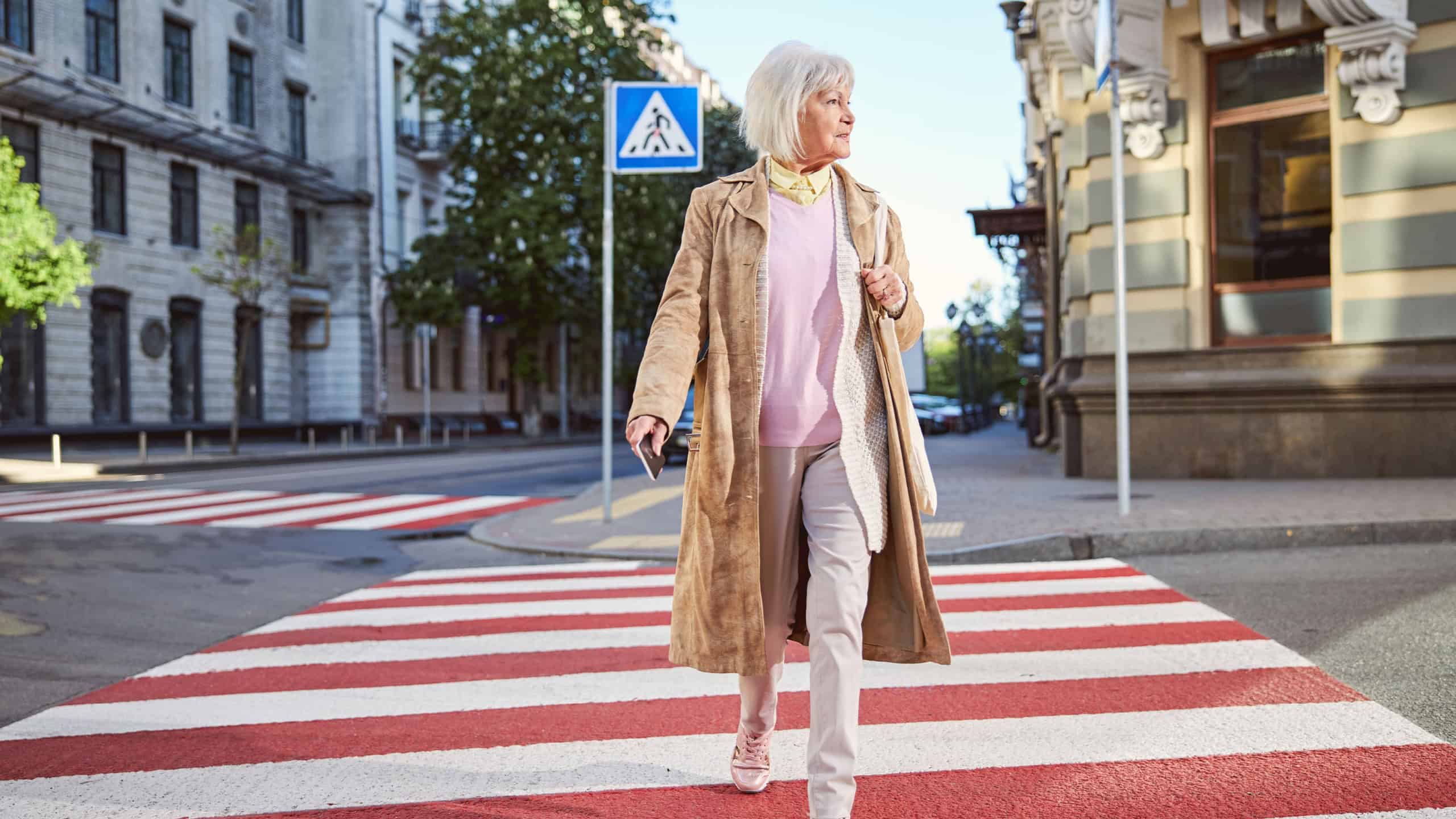 Full length elderly happy female going on the pedestrian crossing to the shopping mall with mobile in hand