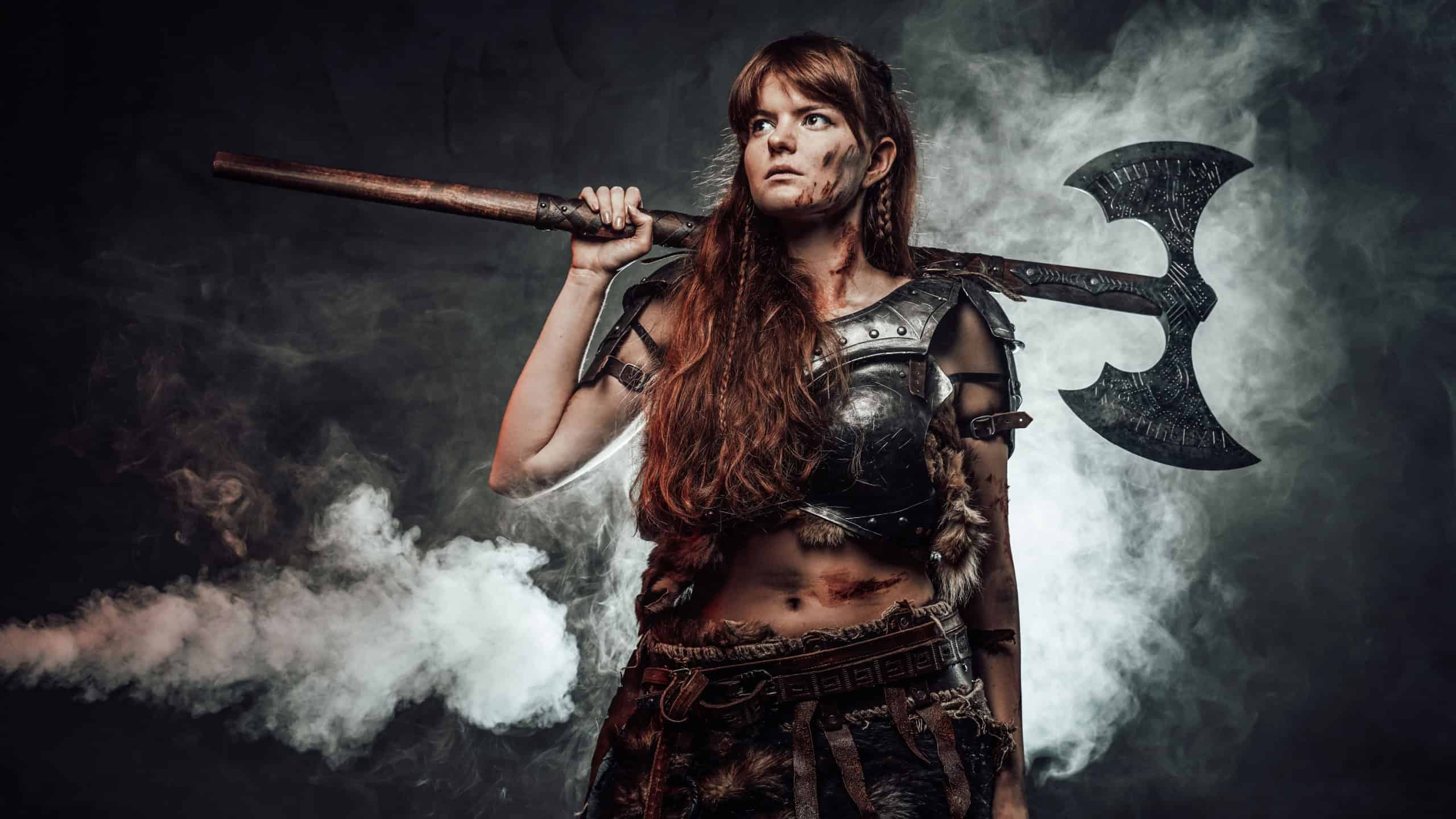 Portrait of woman viking wielding two handed axe and dressed in dark light armour in dark foggy background.