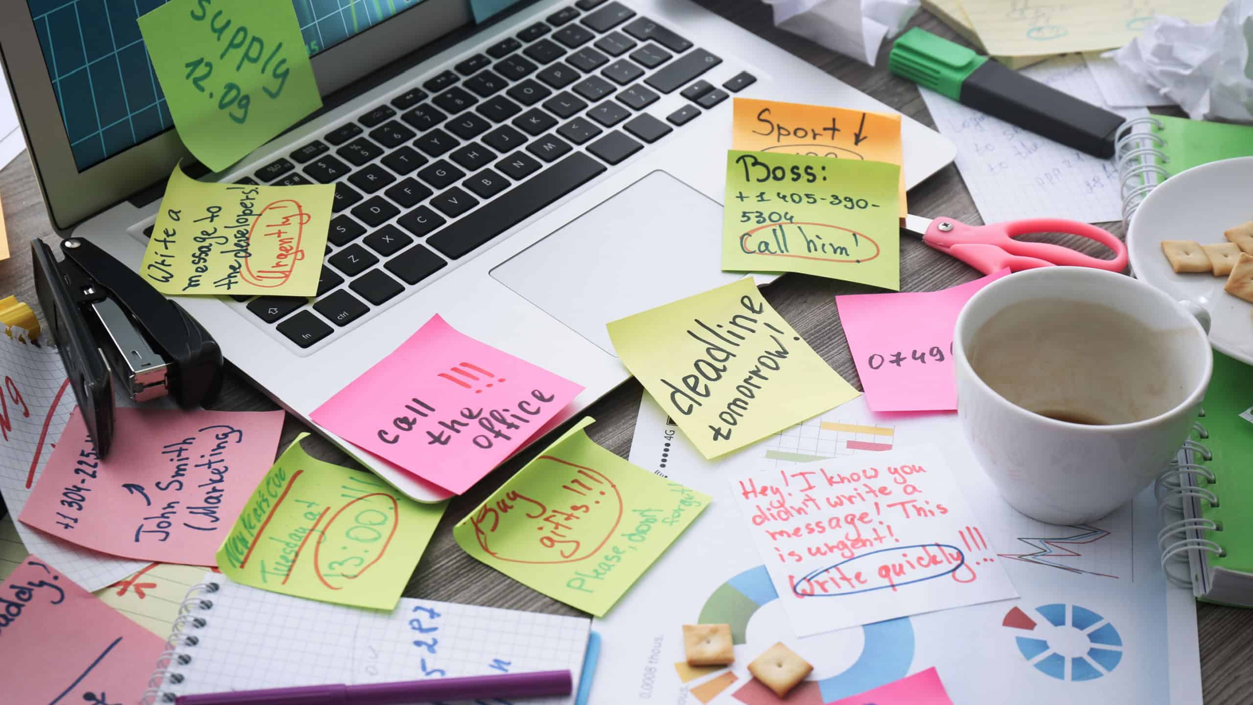 Laptop, notes and office stationery in mess on desk. Overwhelmed with work
