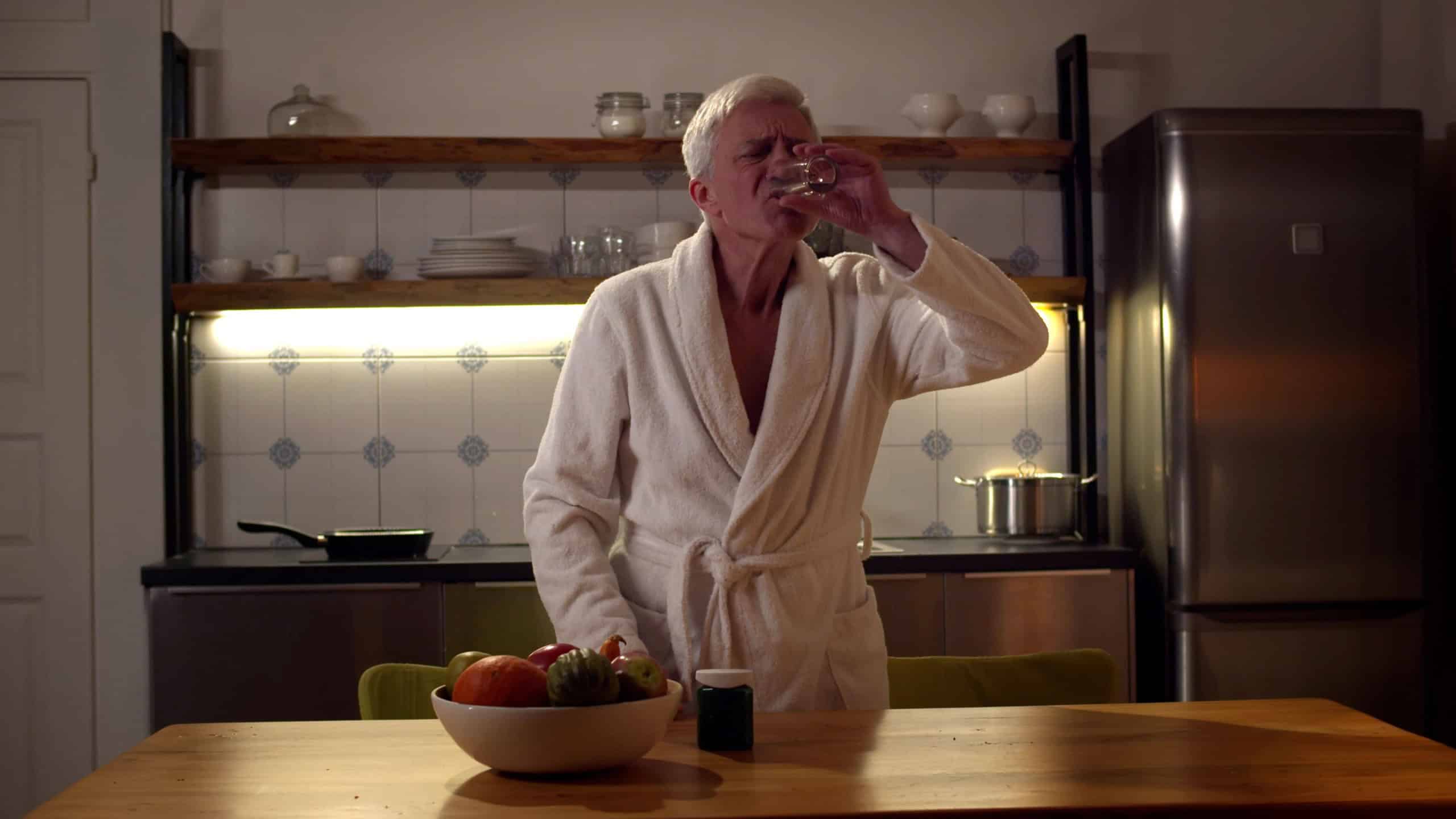 Senior man in bathrobe taking pills and drinking water in kitchen in evening. Aged male suffering from insomnia taking sleeping pills at night. Mature man taking medication in kitchen