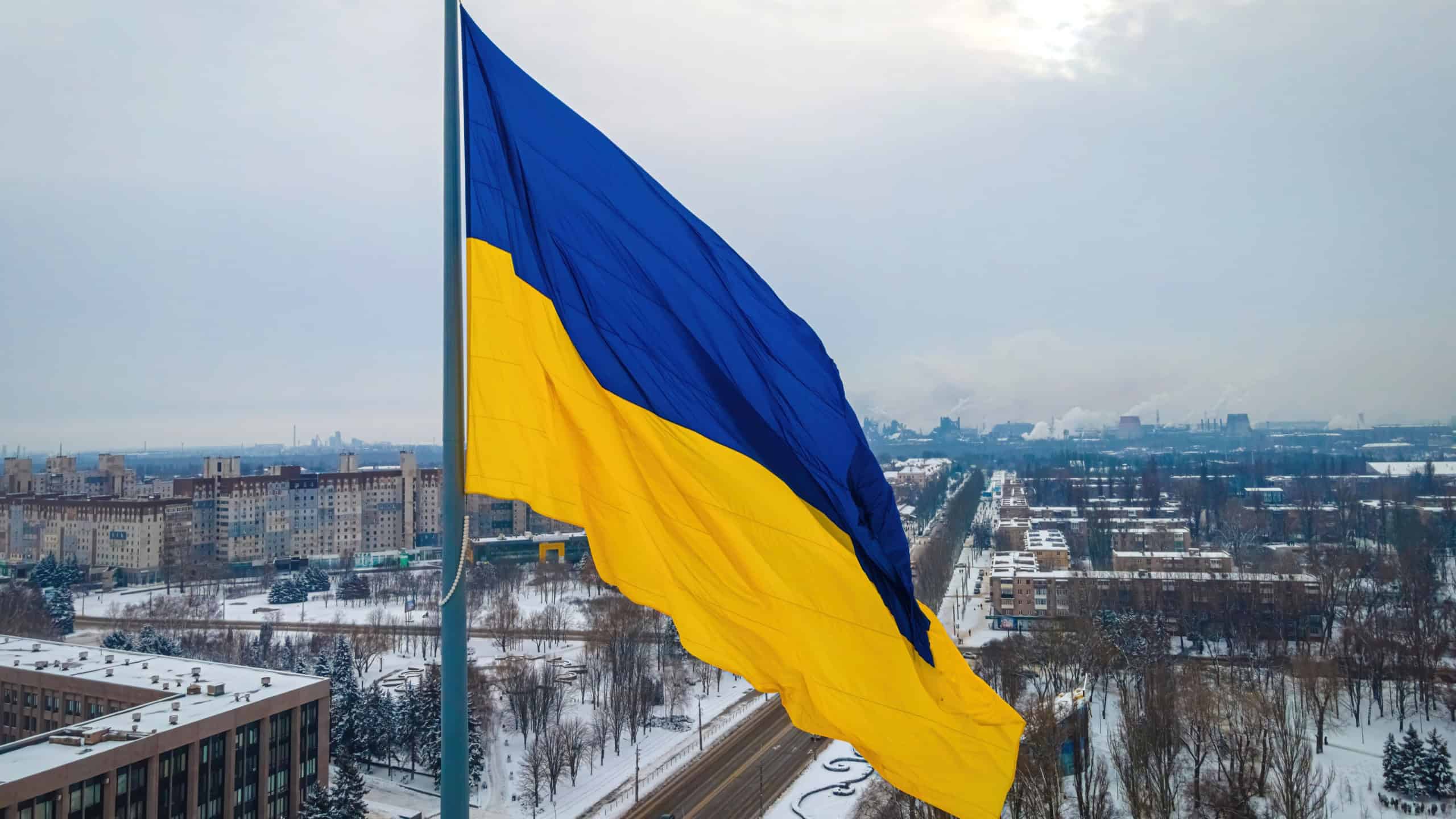 Large Ukraine flag waving above the city in winter light, symbol of national pride and resilience during war; perfect for news, politics, charity, solidarity and freedom themes