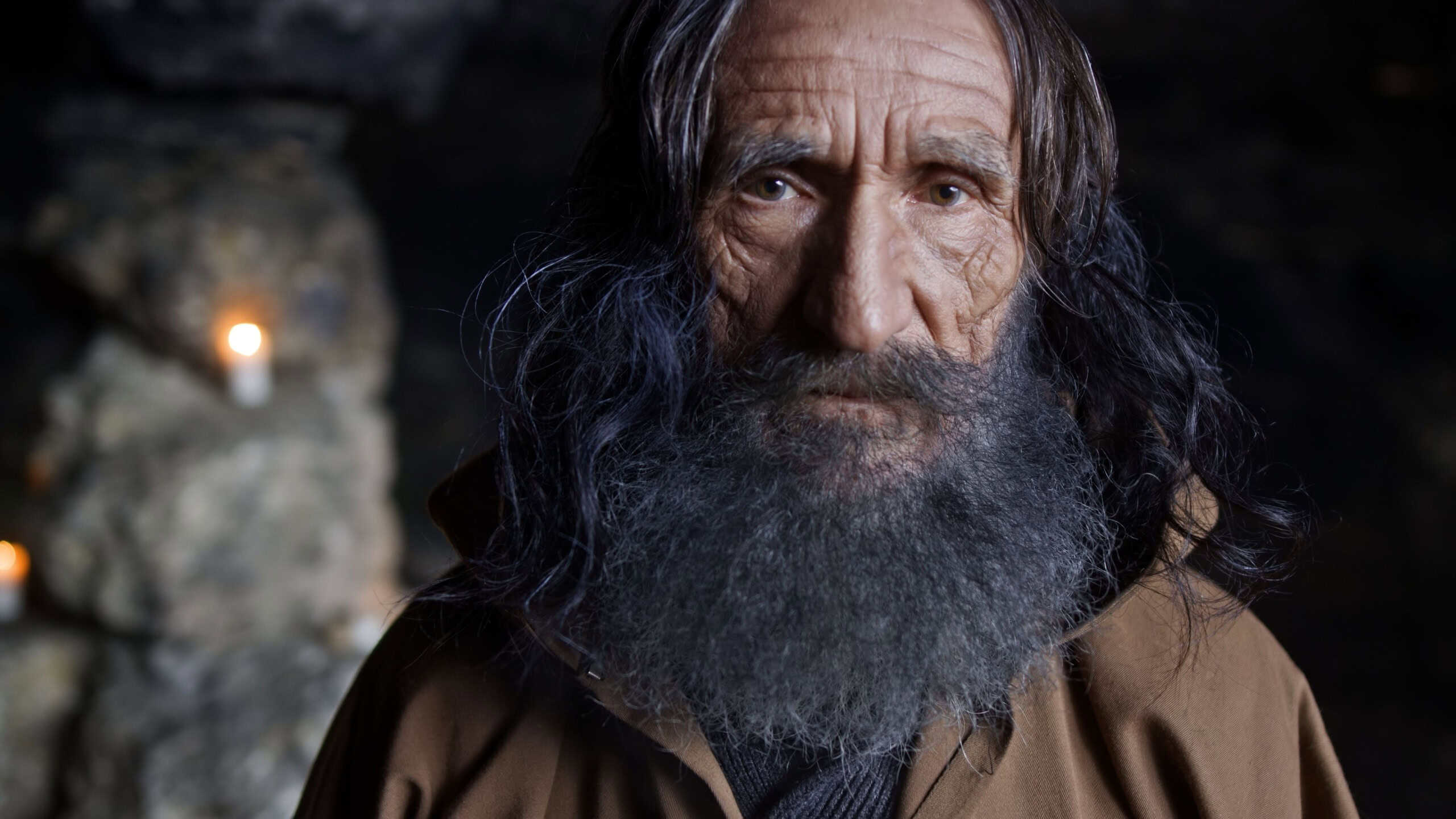 Portrait of senior sage man in cassock and with gray hair standing in rocky cave with candles and looking at camera