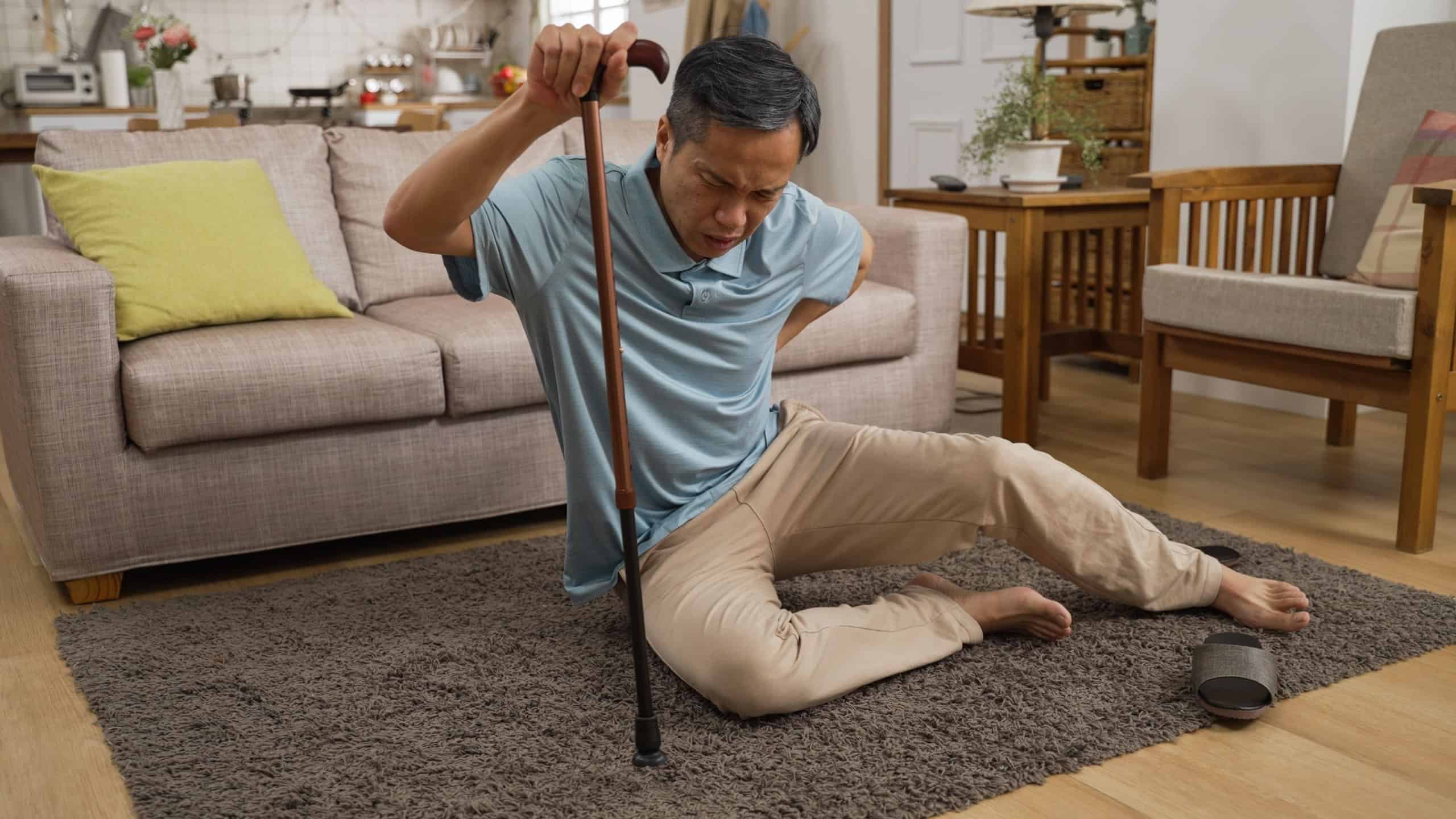 Asian injured mature male groaning in pain while he tries to get up with a stick after fall at home. He holds his lower back and can barely stand up