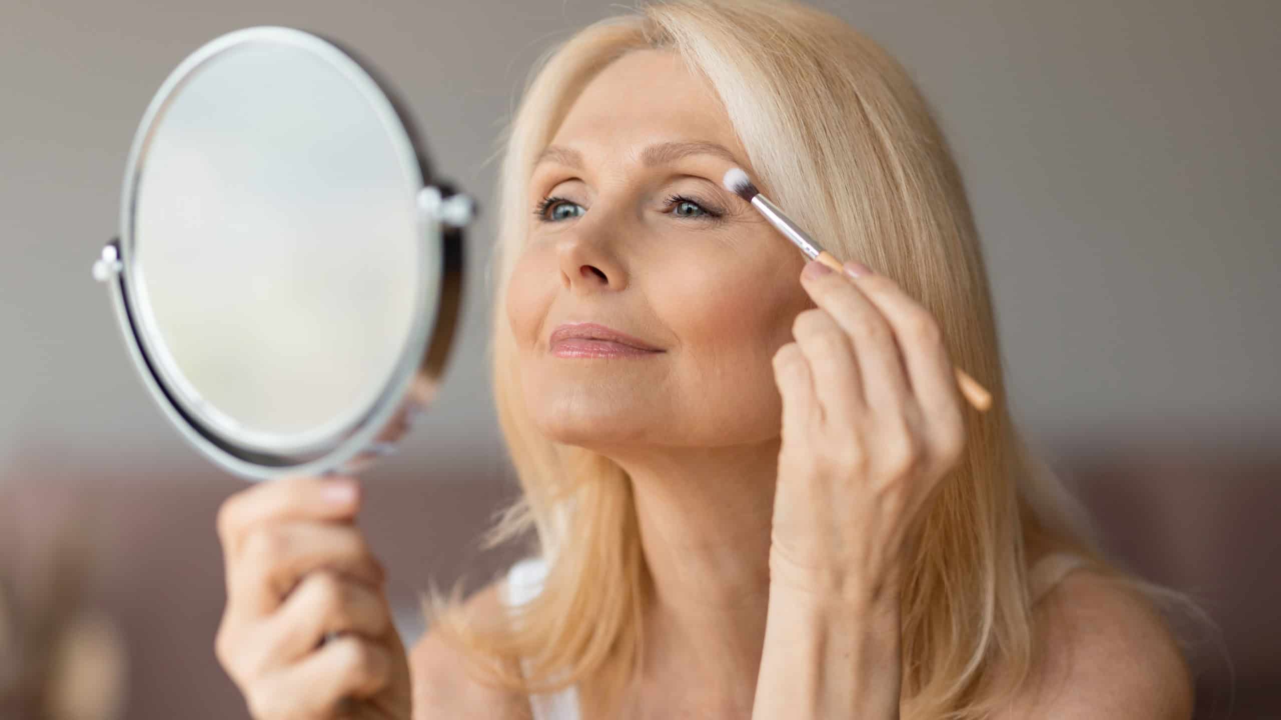 Happy senior european woman applying eyeshadow with brush, looking in mirror and smiling, female doing nude makeup. Anti-aging cosmetics, beauty care