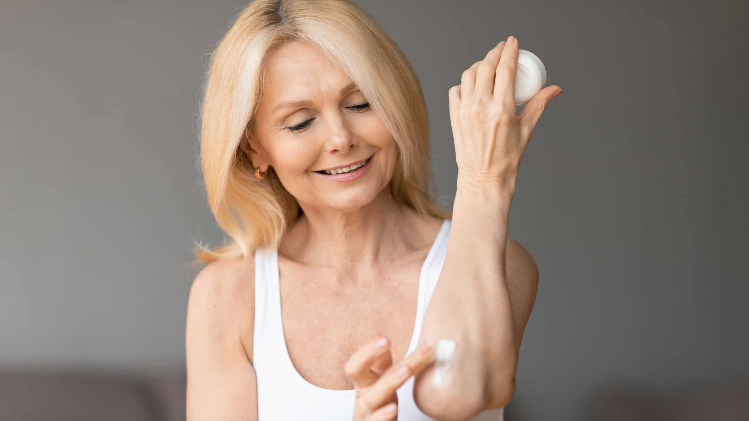 Body care concept. Happy aged woman rubbing moisturising lotion on elbow while pampering herself at home, female applying nourishing cream, copy space