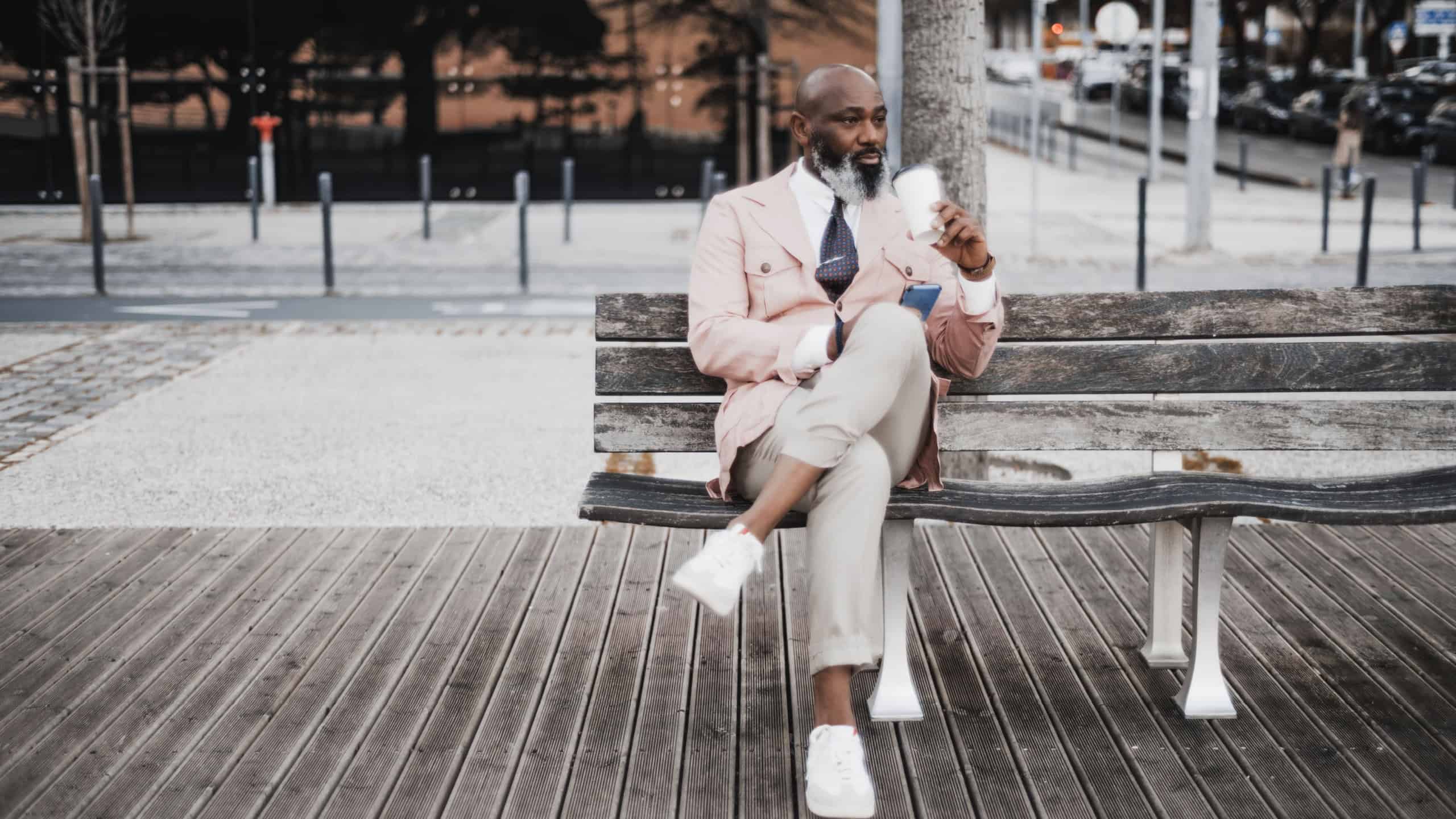 Vertical shot: A well-dressed bald black man seated on a wooden bench with a coffee cup nearby. His peachy jacket, cream trousers, and phone in hand reflect his confident and stylish demeanor