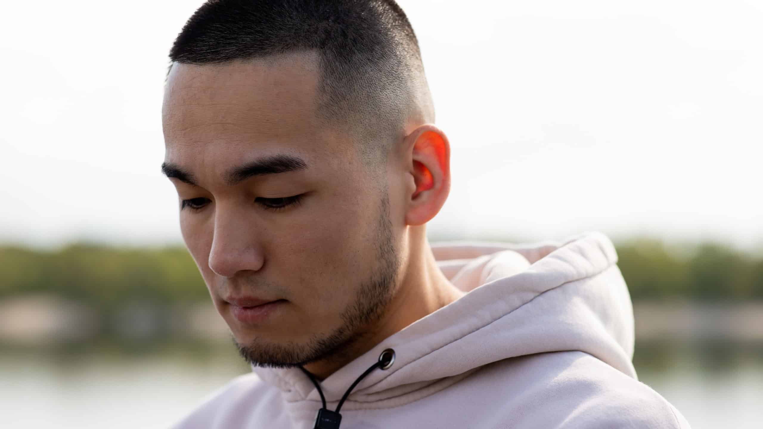 An Asian guy with stubble is standing in front of the camera and looking down. Street photograph of a young man standing in front of a blurred river.