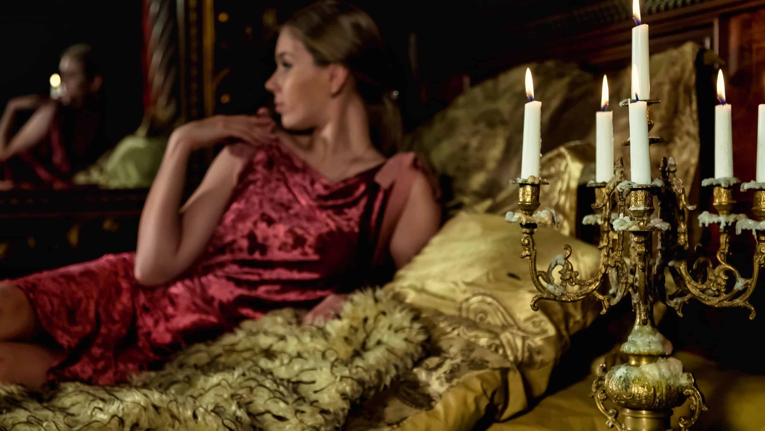 Lovely woman in stylish image posing lying on bed in medieval bedroom, selective focus an candlestick. Pensive young lady in interior antique gold room. Concept of theatre perform. Copy ad text space