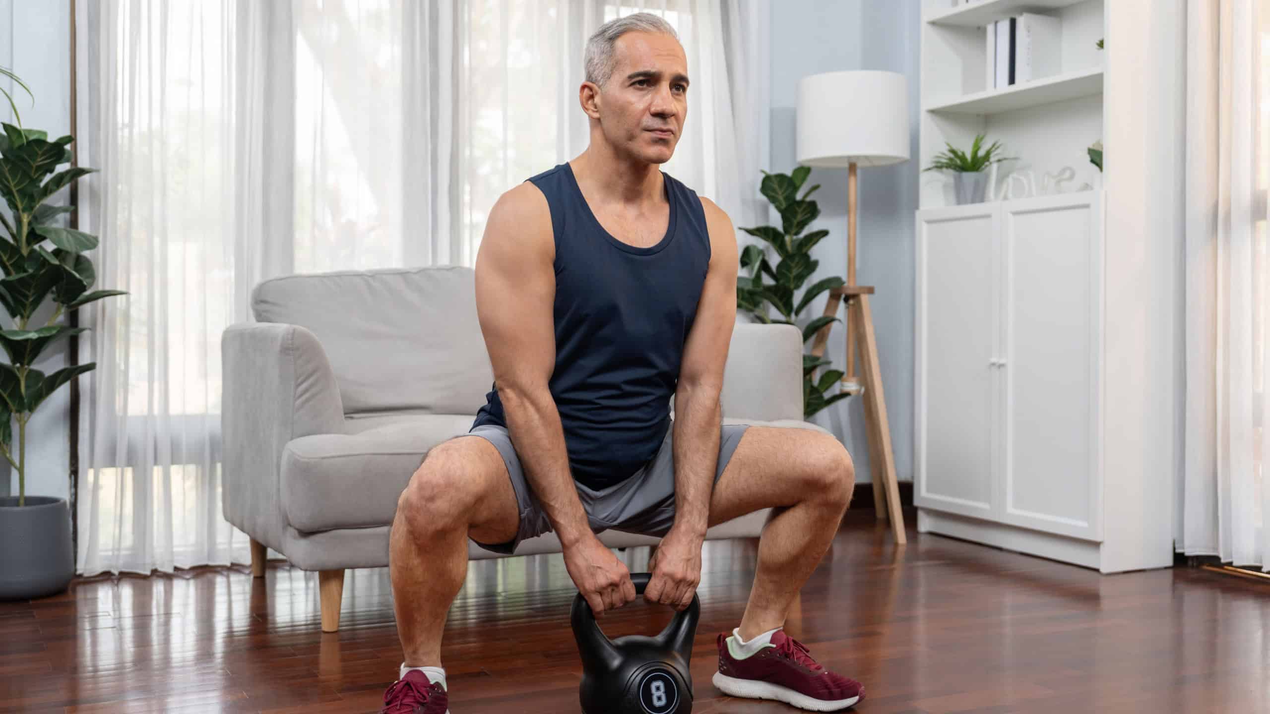 Athletic and sporty senior man engaging in leg day training session with squat and bodyweight kettle ball at home exercise as concept of healthy fit body lifestyle after retirement. Clout