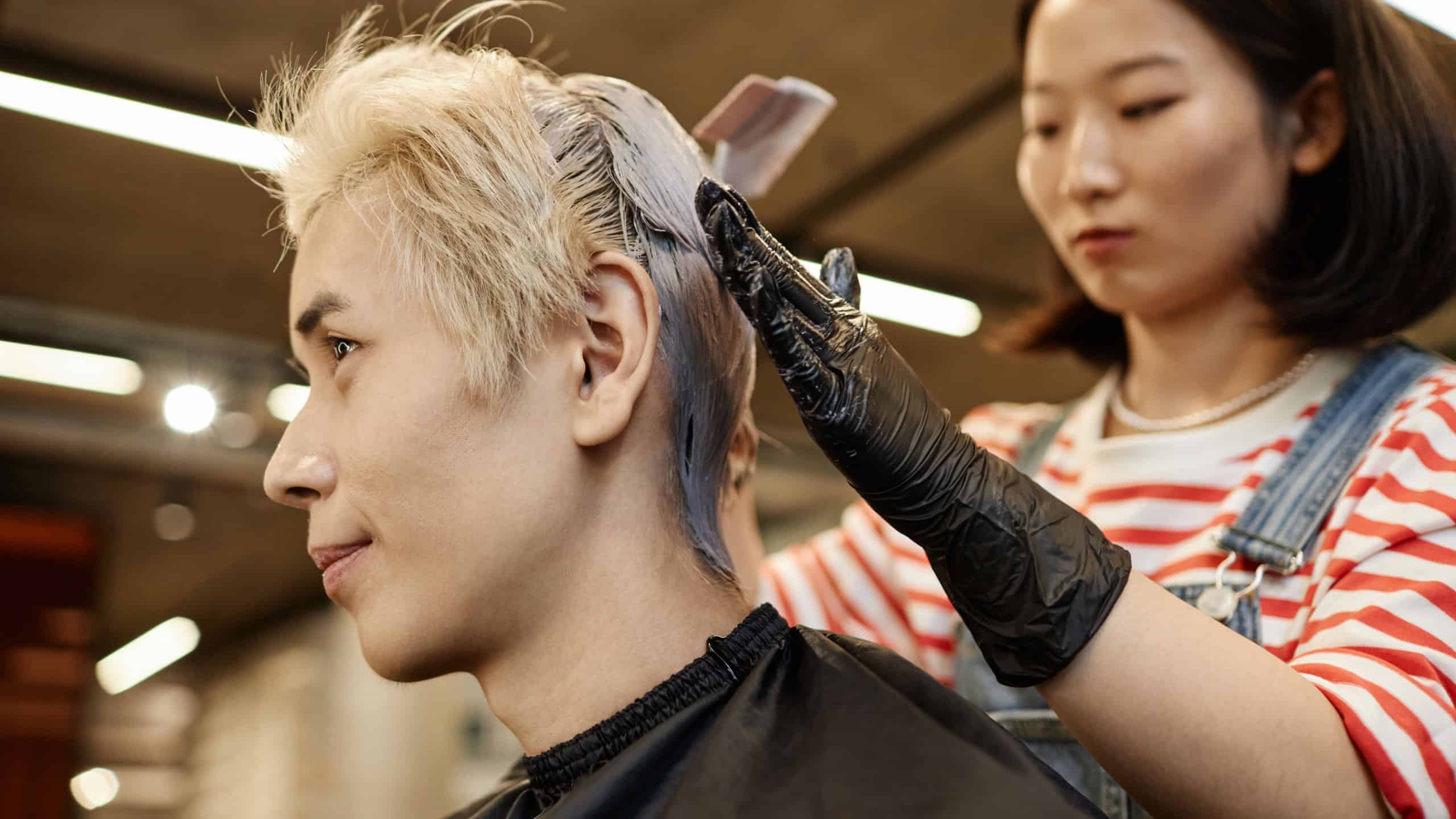Side view portrait of young Asian man in beauty salon with hairstylist bleaching or dyeing hair