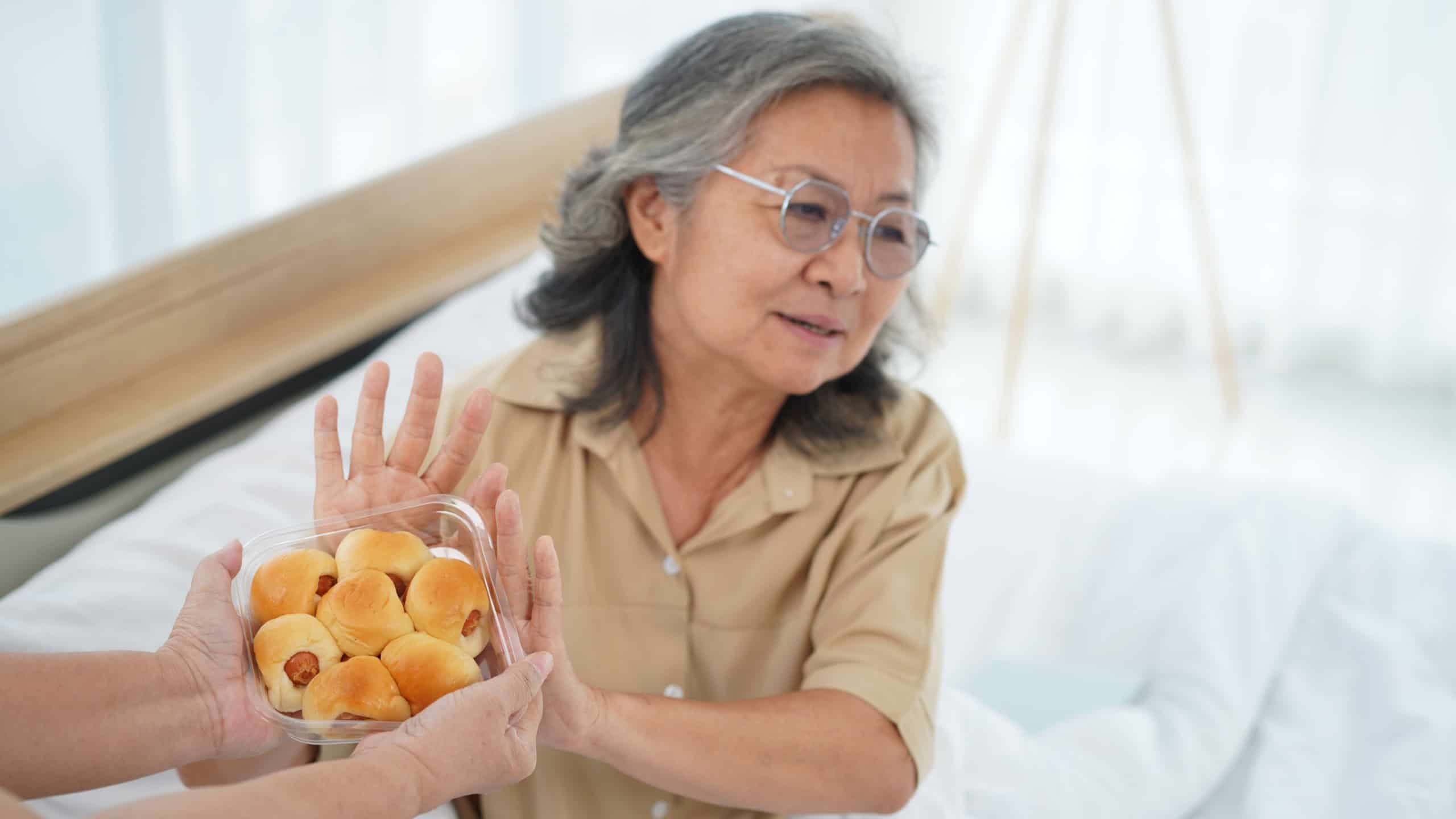 High angle view. Selective focus. Senior Asian woman wearing glasses sitting on bed. She raised hand and turned away, refusing to accept the bread from the caregiver.